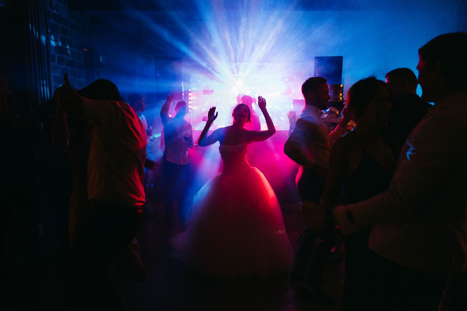 Bride in white ball gown dancing under colorful stage lights with guests at a lively wedding reception party.