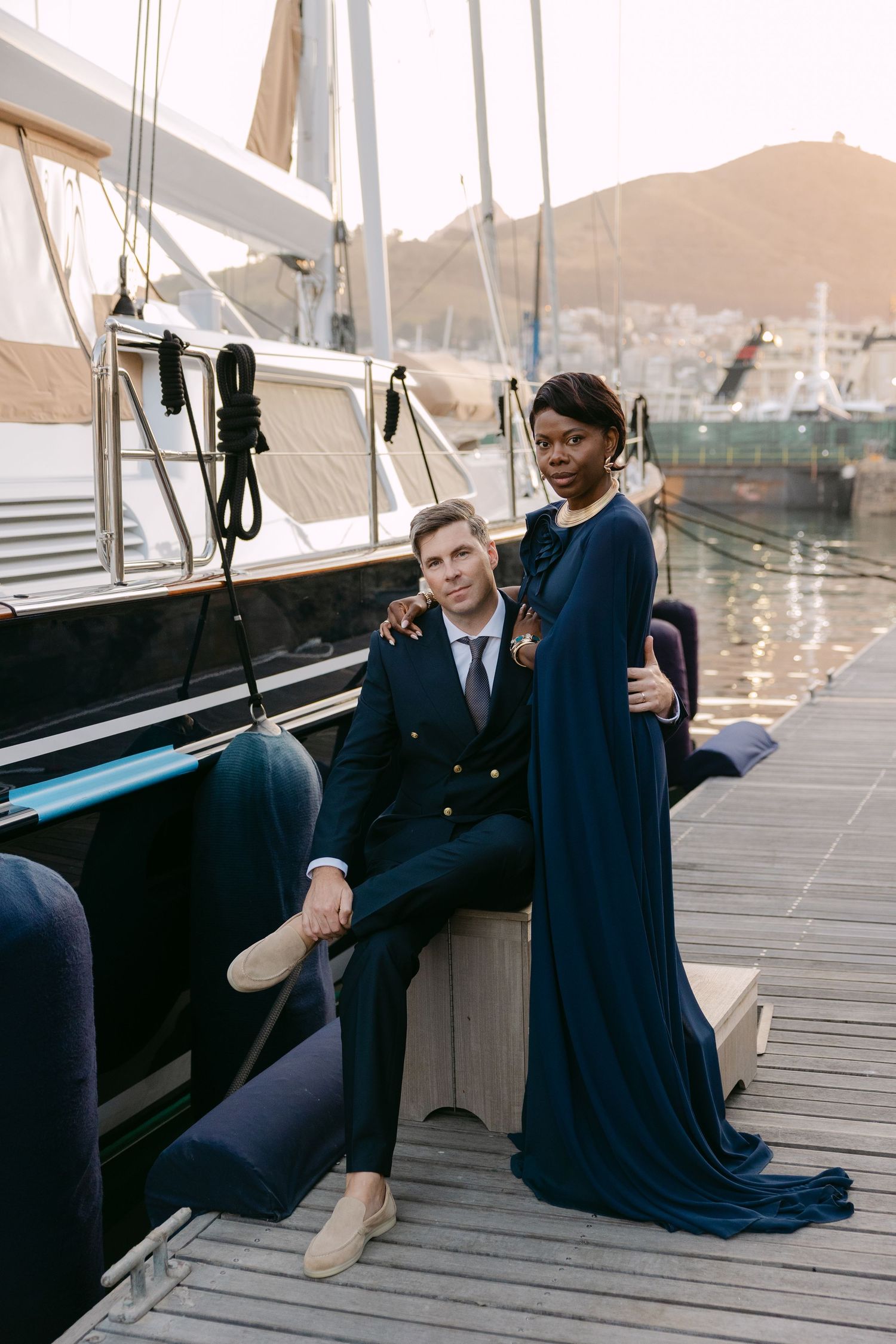 A couple in navy blue formal attire sits together on a wooden dock by the water at sunset.