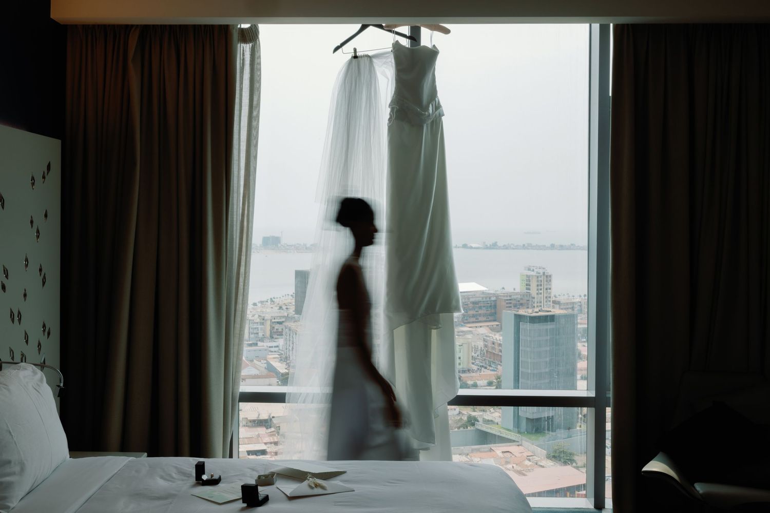 Bride silhouette standing by hotel window with wedding dress hanging, overlooking city skyline and waterfront view.