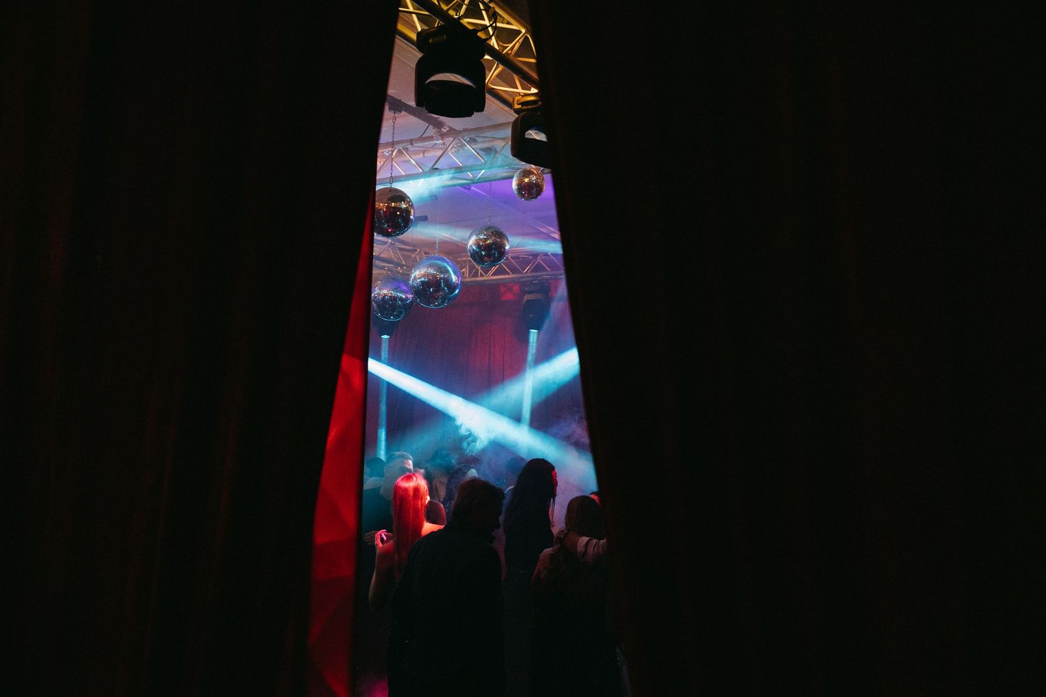Nightclub dance floor with disco balls, colorful stage lights, and silhouetted crowd visible through dark curtains.