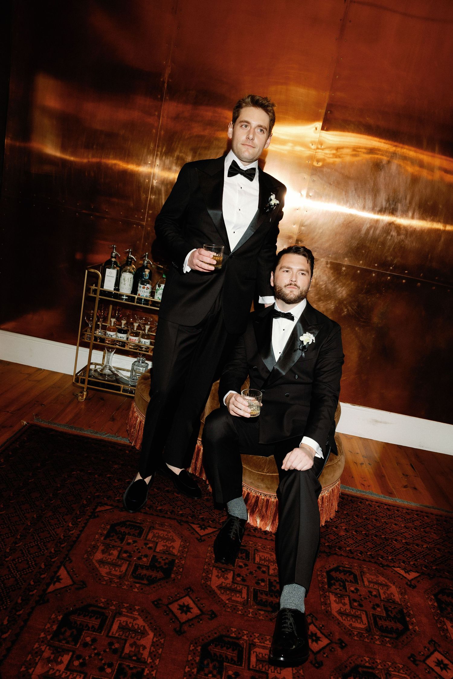 Two people in formal black tuxedos pose on a patterned carpet in a dimly lit room with warm lighting.