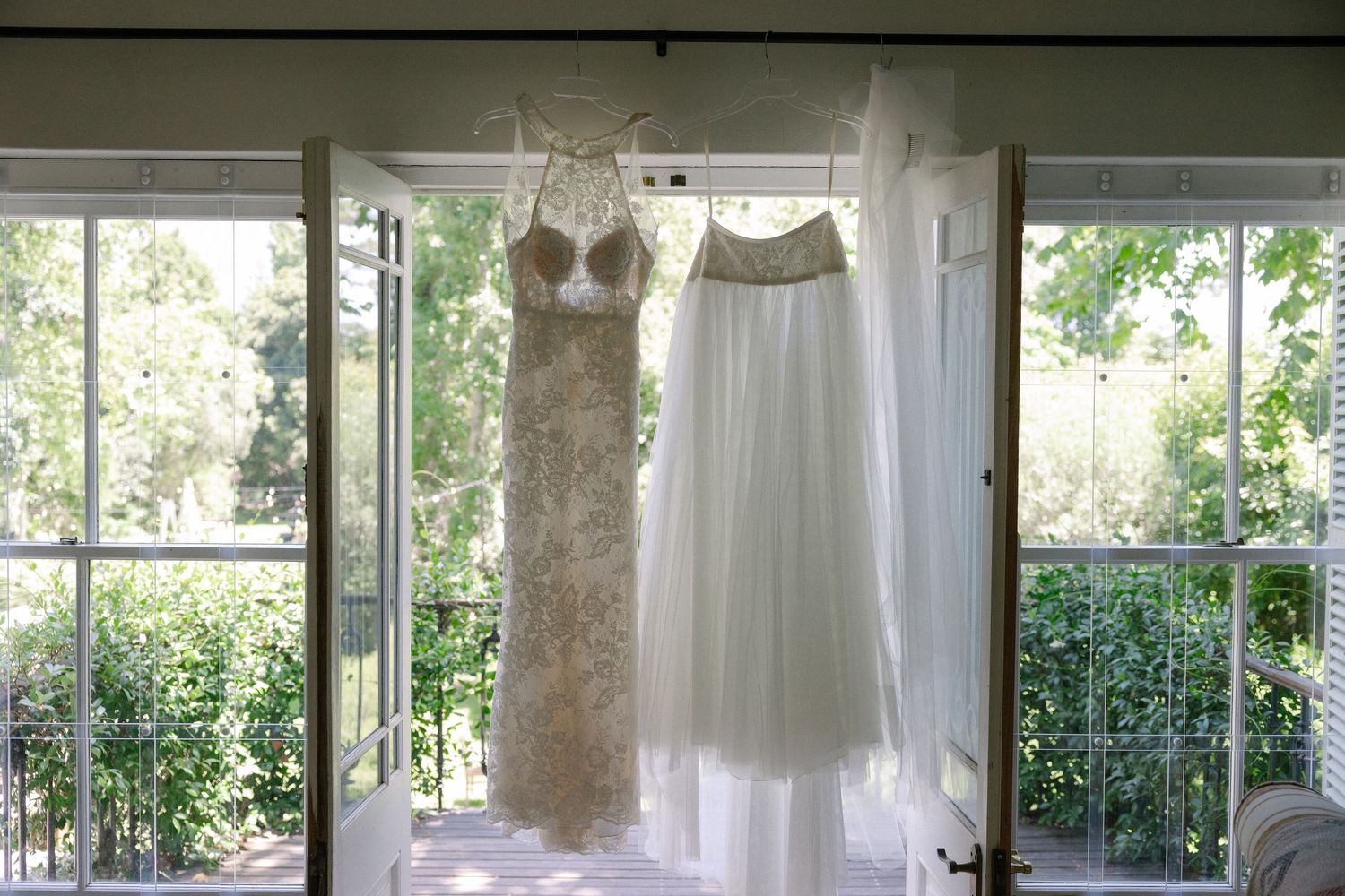 Two elegant wedding dresses hanging on a door frame with lush green trees visible through large windows.