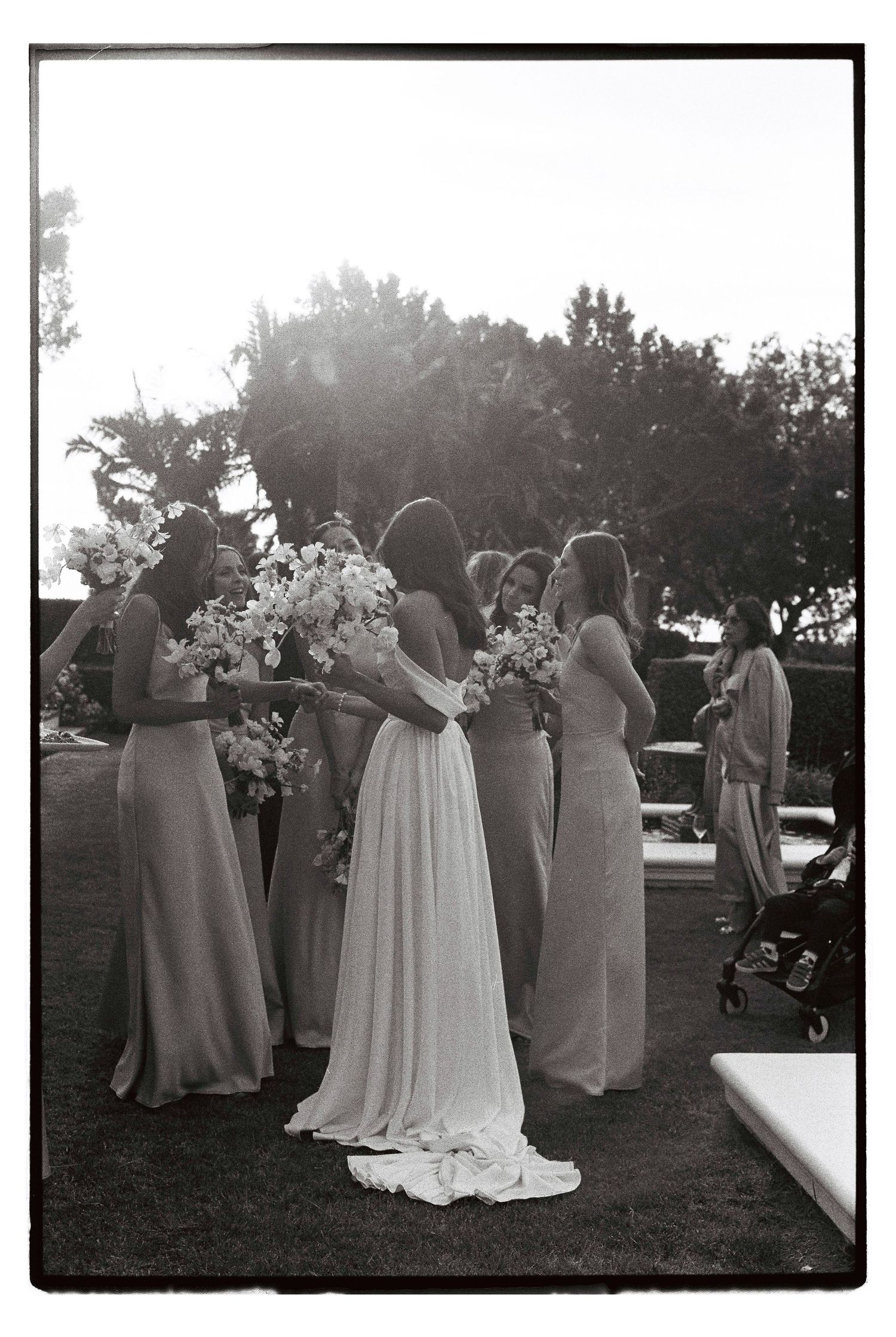 A wedding party gathers outdoors with bouquets of white flowers during a sunlit celebration.