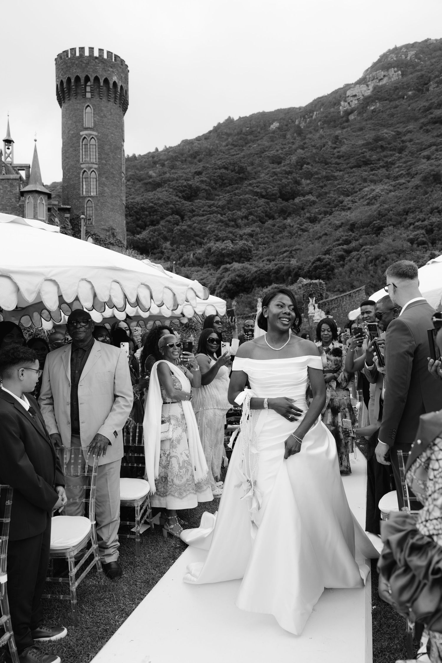 Bride walks down the aisle in elegant white gown at outdoor castle wedding ceremony with guests watching.