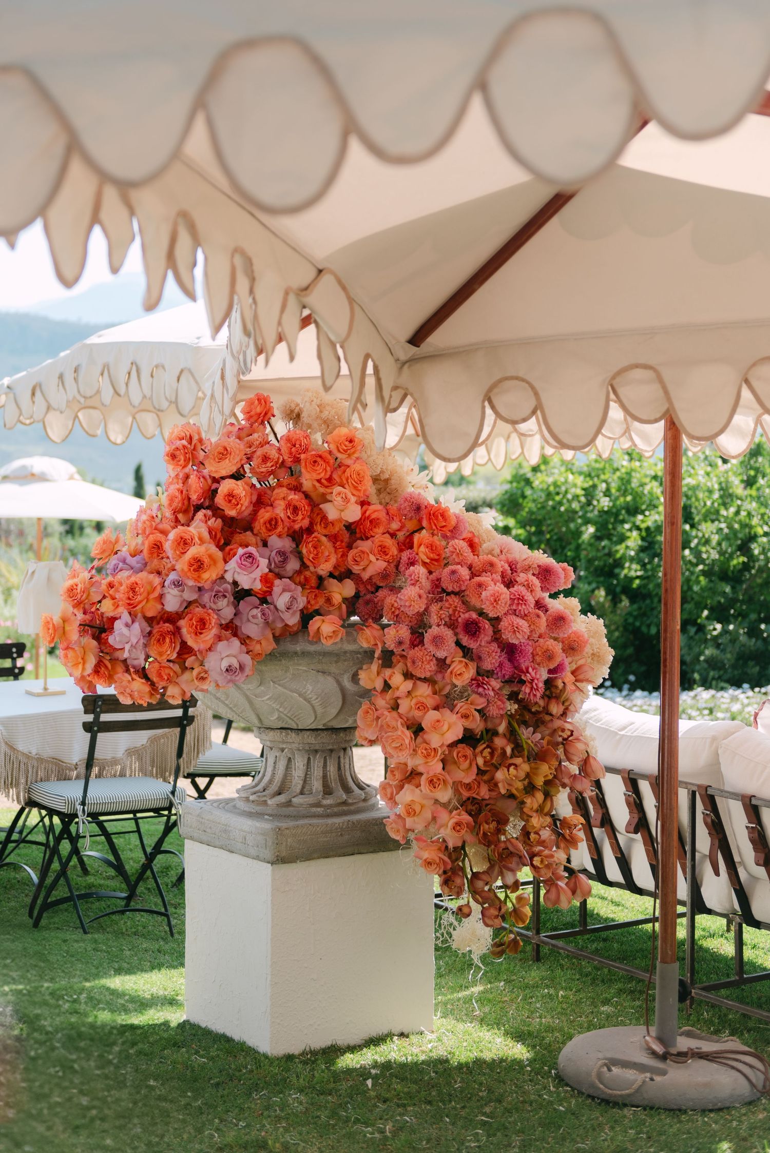 Lush orange and pink floral arrangement in stone urn under elegant cream scalloped tent at outdoor wedding reception.
