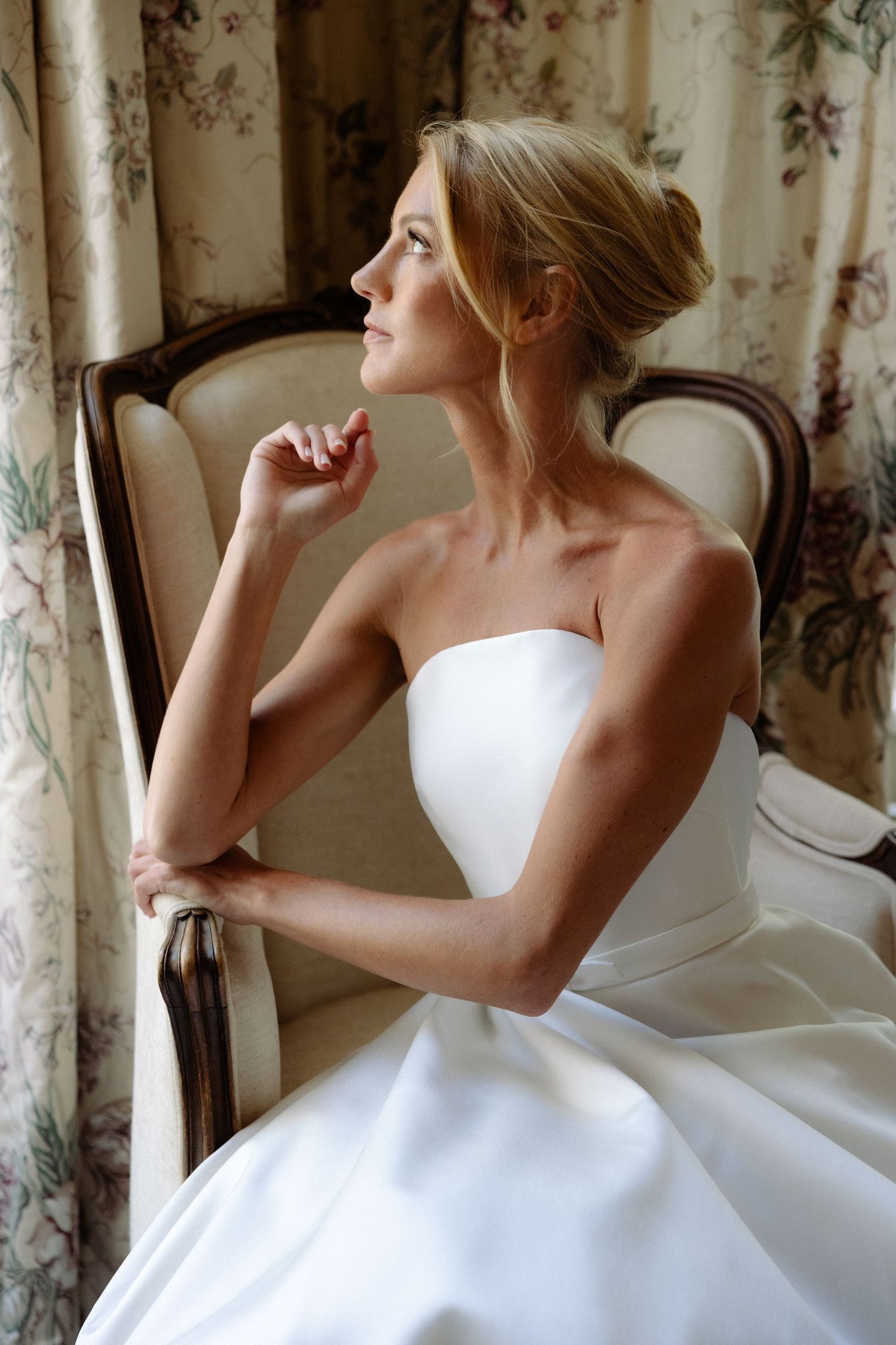 A bride in a white strapless gown gazes thoughtfully out a window with floral curtains in an elegant room.