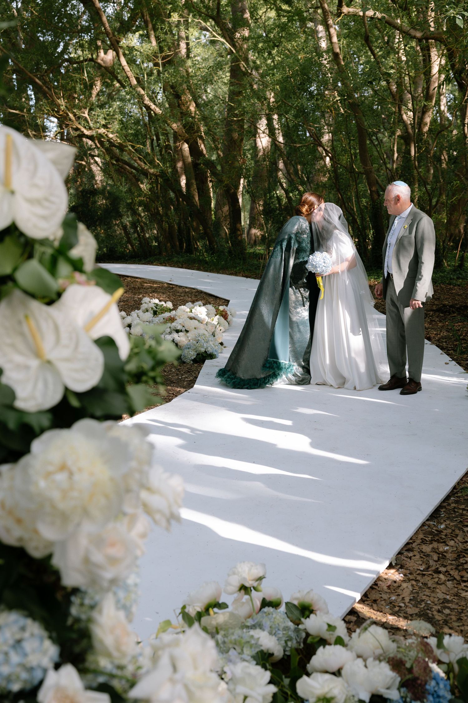 Outdoor wedding ceremony setup in a wooded area with white floral decorations and a white aisle runner.