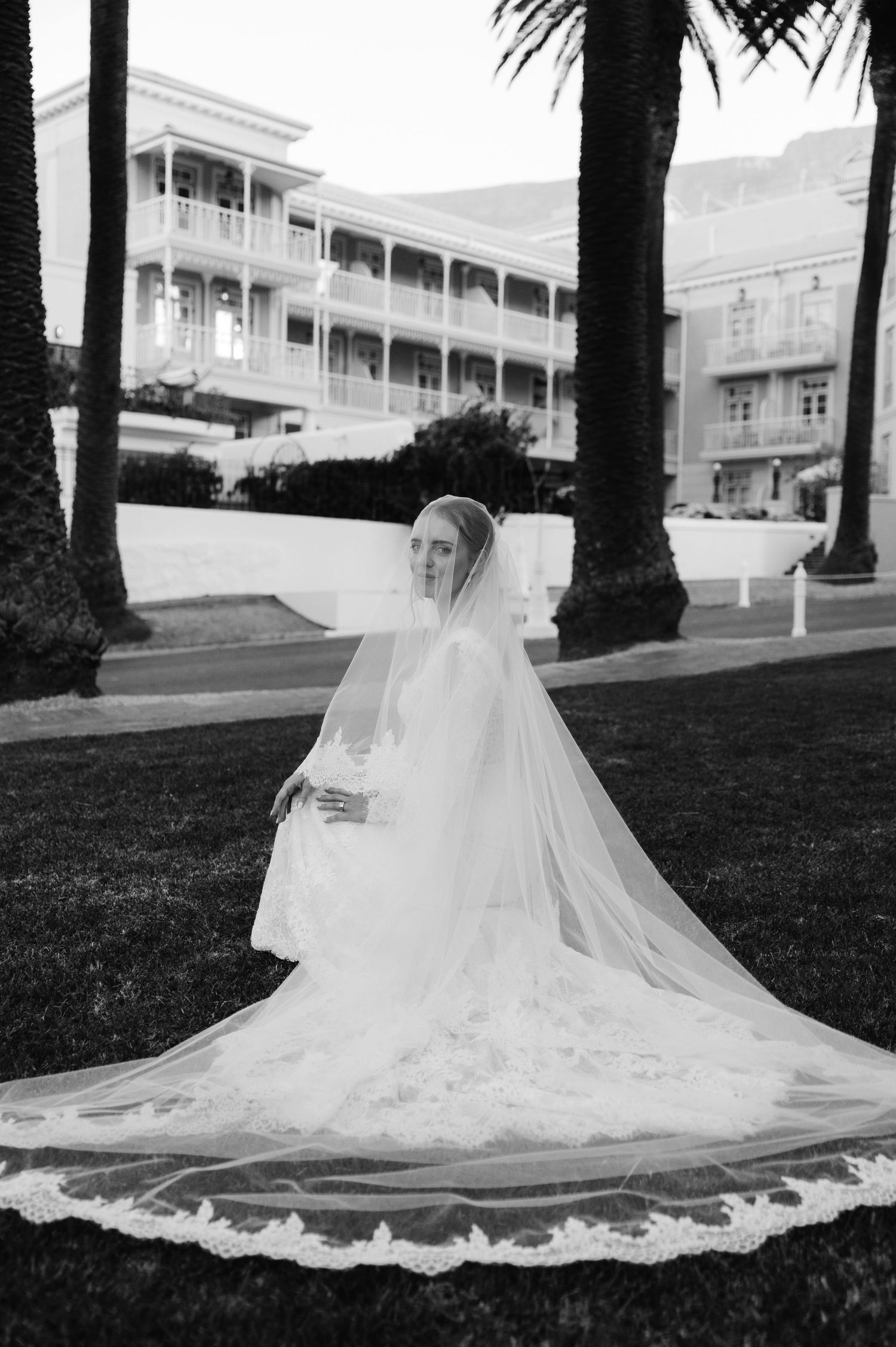 A flowing white wedding dress with a long cathedral veil spread across a manicured lawn in front of a grand building.