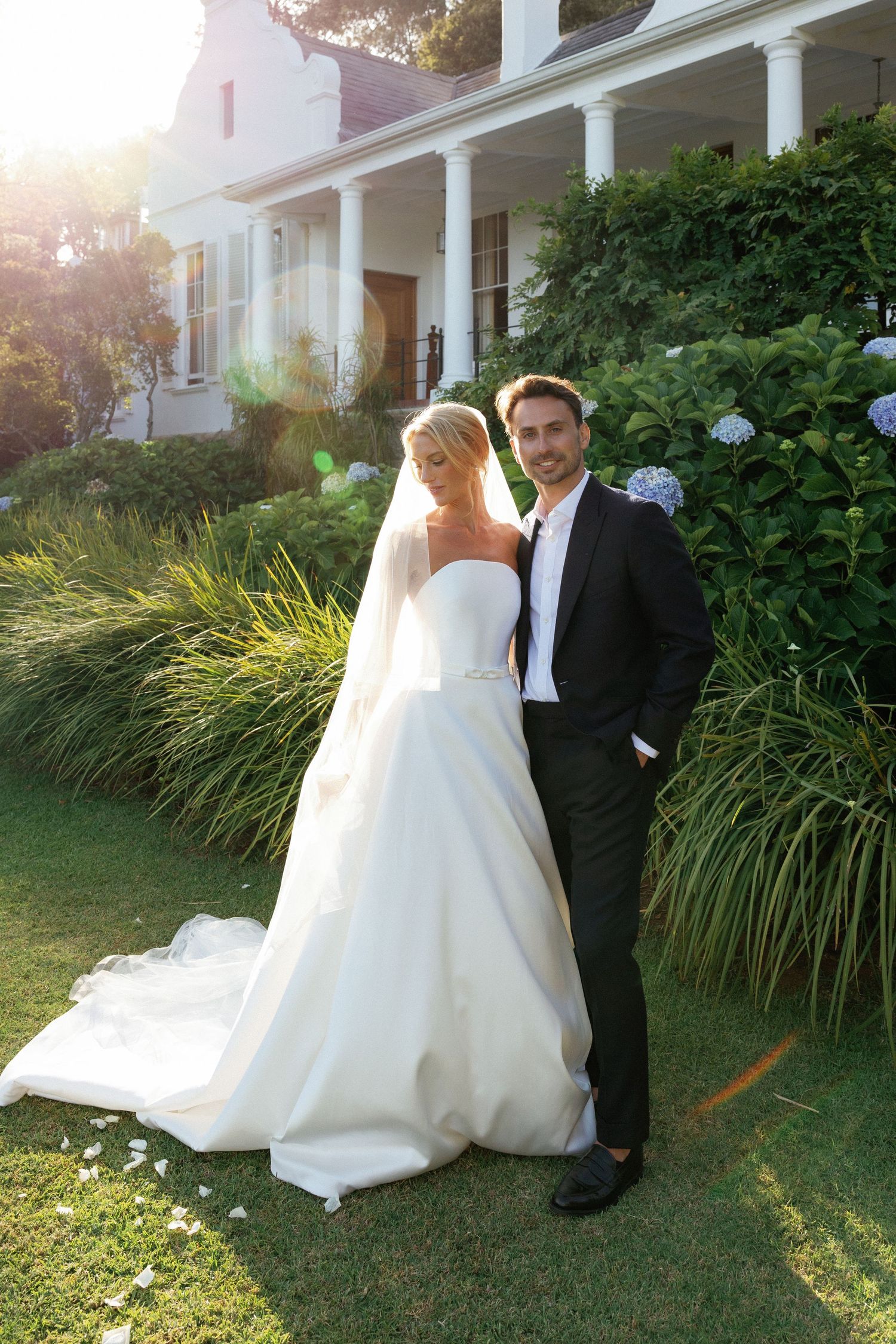 Bride in white gown and groom in black tuxedo pose together on lawn outside elegant white colonial mansion.