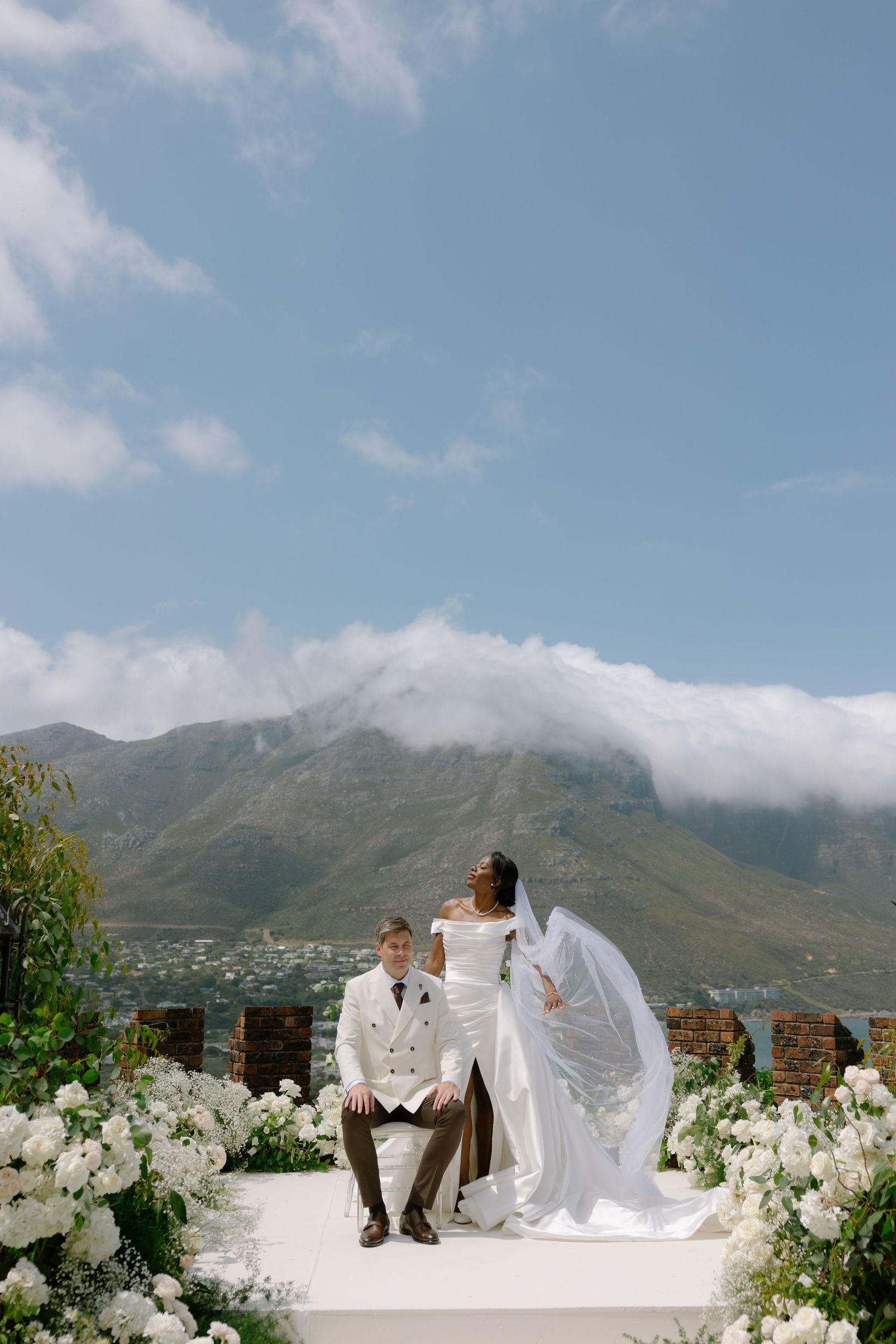 A romantic wedding couple sits on a white wall overlooking mountains and ocean in Cape Town, South Africa.
