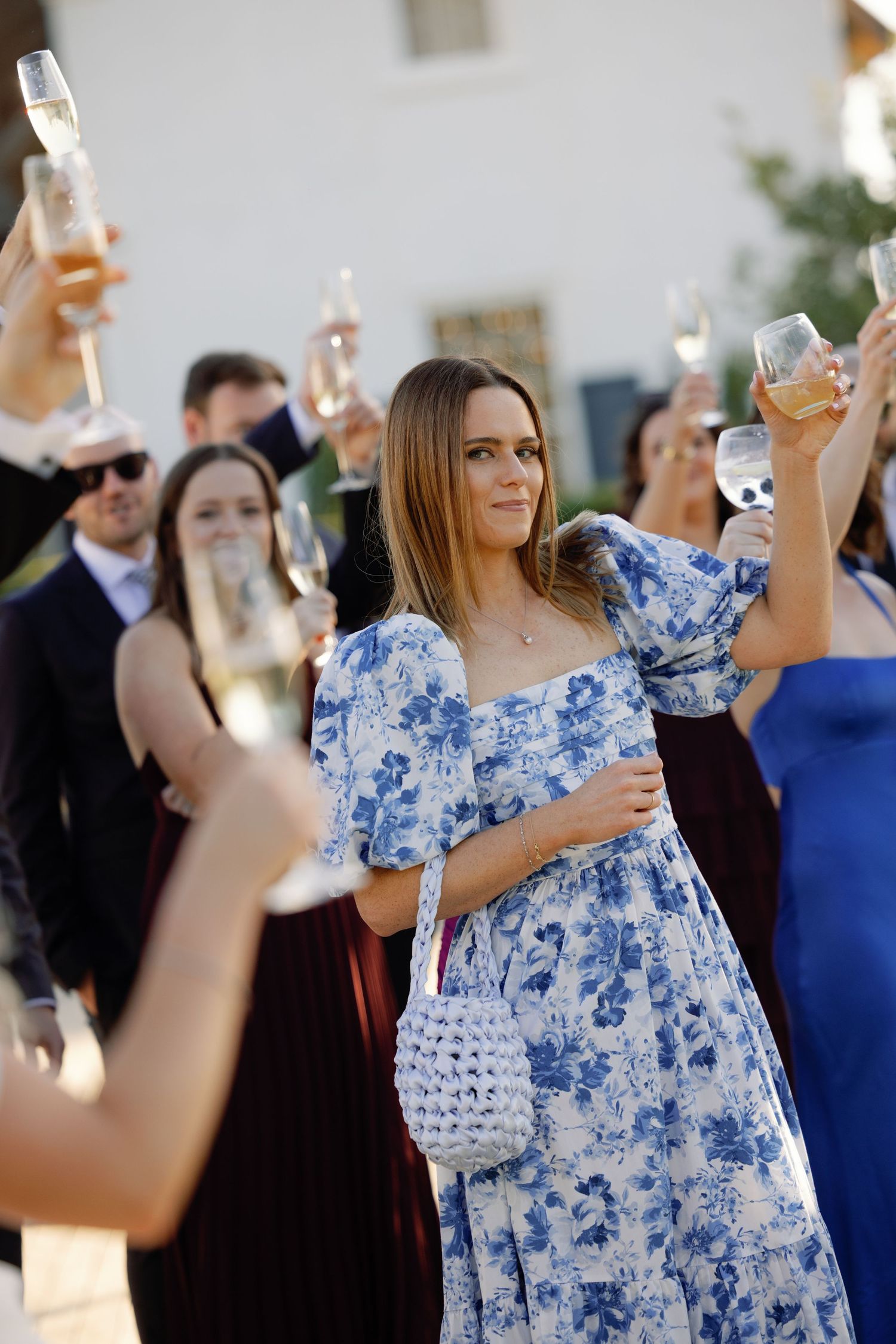 Woman in blue floral dress raising champagne glass at outdoor wedding celebration with guests.