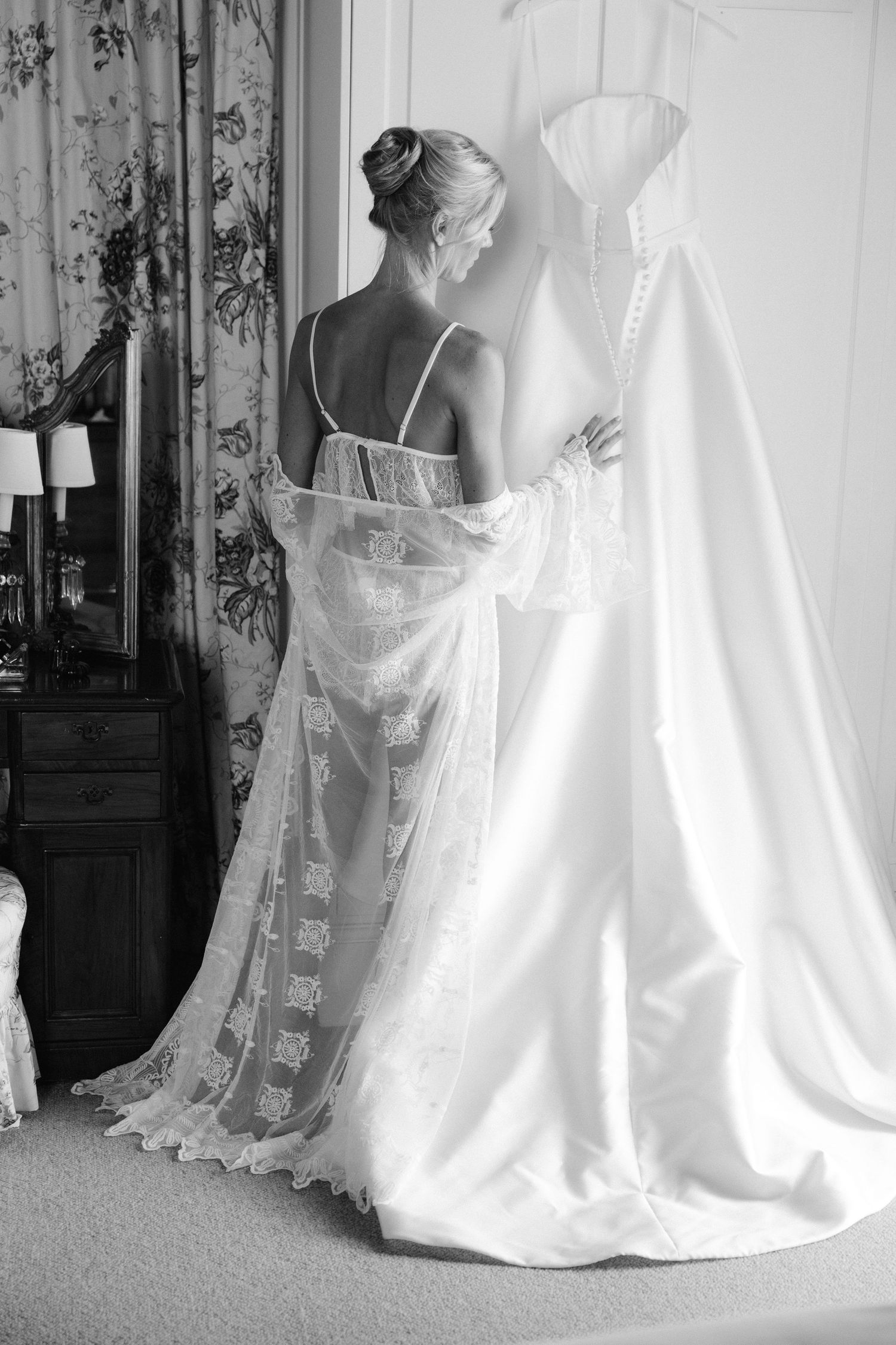Bride in lace robe admires her elegant white wedding gown hanging in a sunlit room, black and white photo.