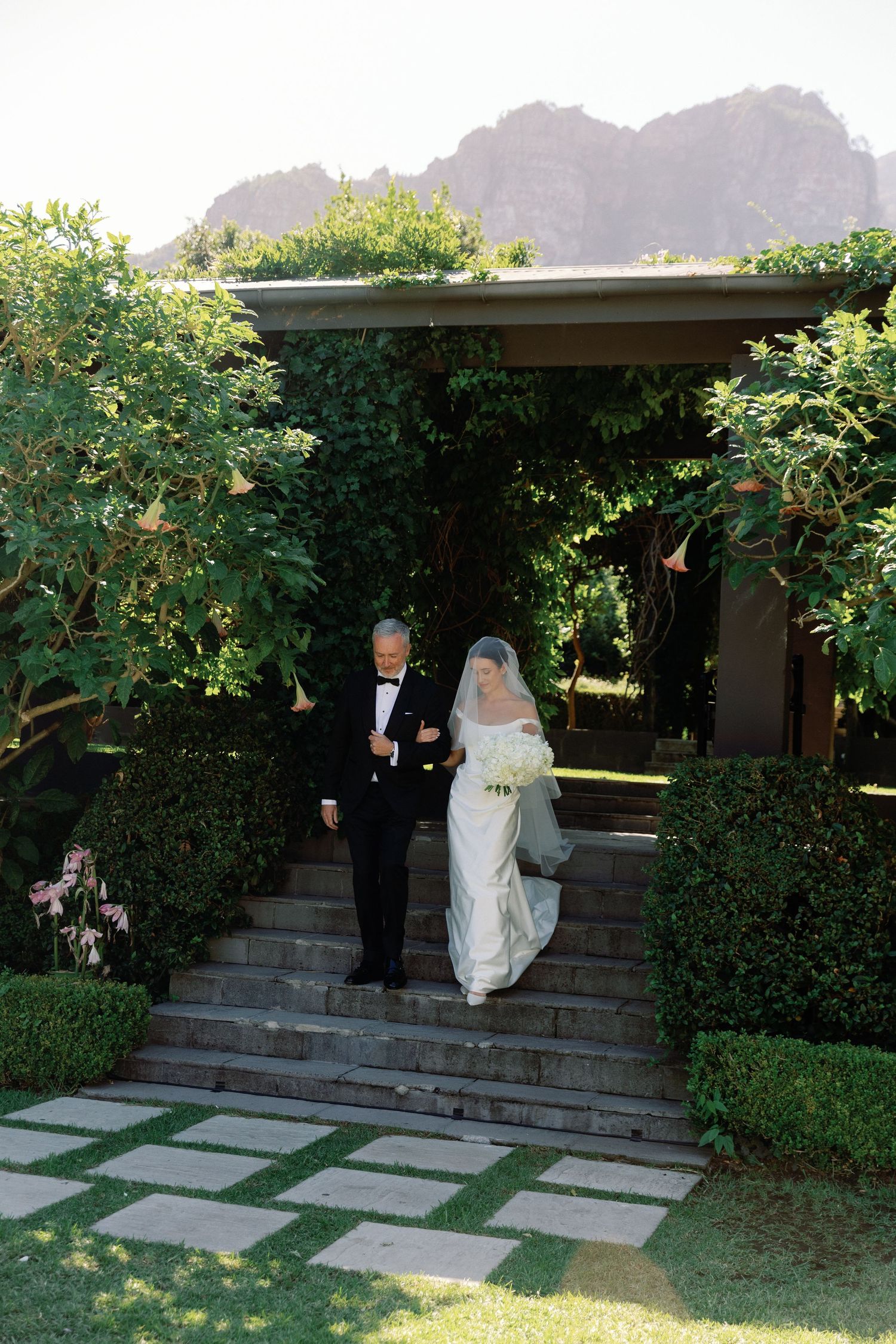Bride in white gown and veil walks with escort down garden steps, lush greenery and mountain backdrop.