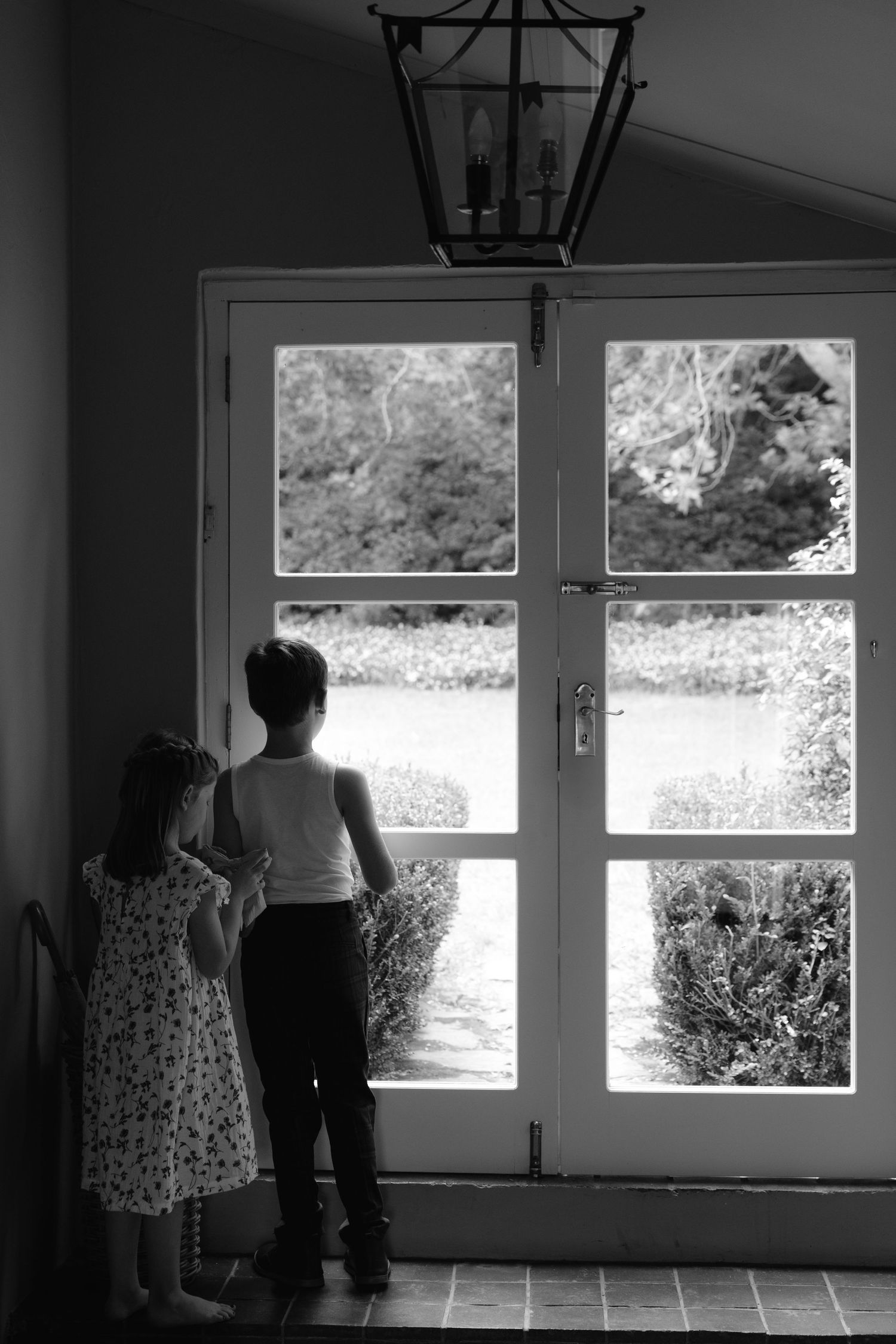 Two figures stand silhouetted against a bright doorway with glass panels and a lantern hanging above in black and white.