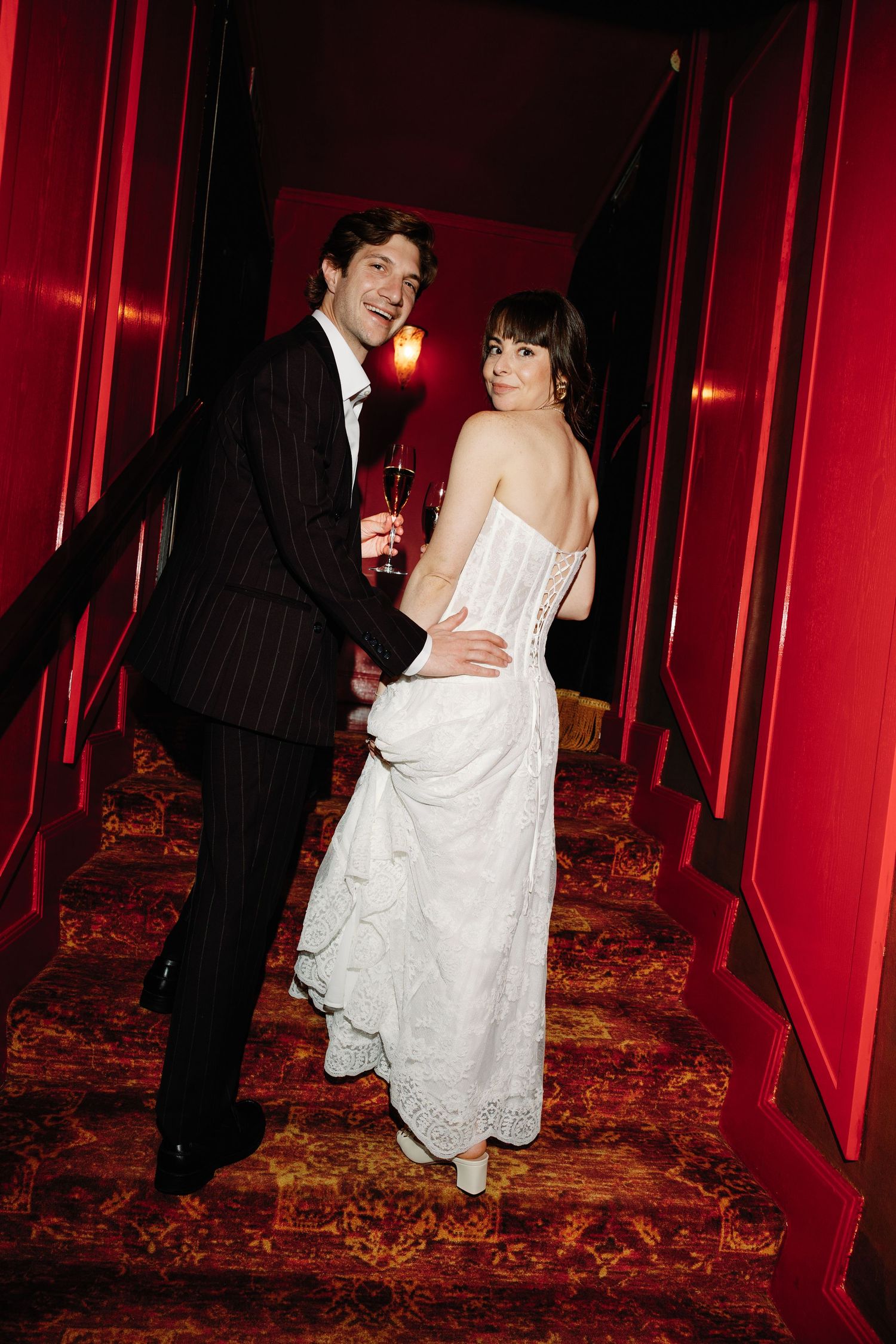 A couple in formal attire playfully poses in a red-walled hallway with ornate carpeting at an event.