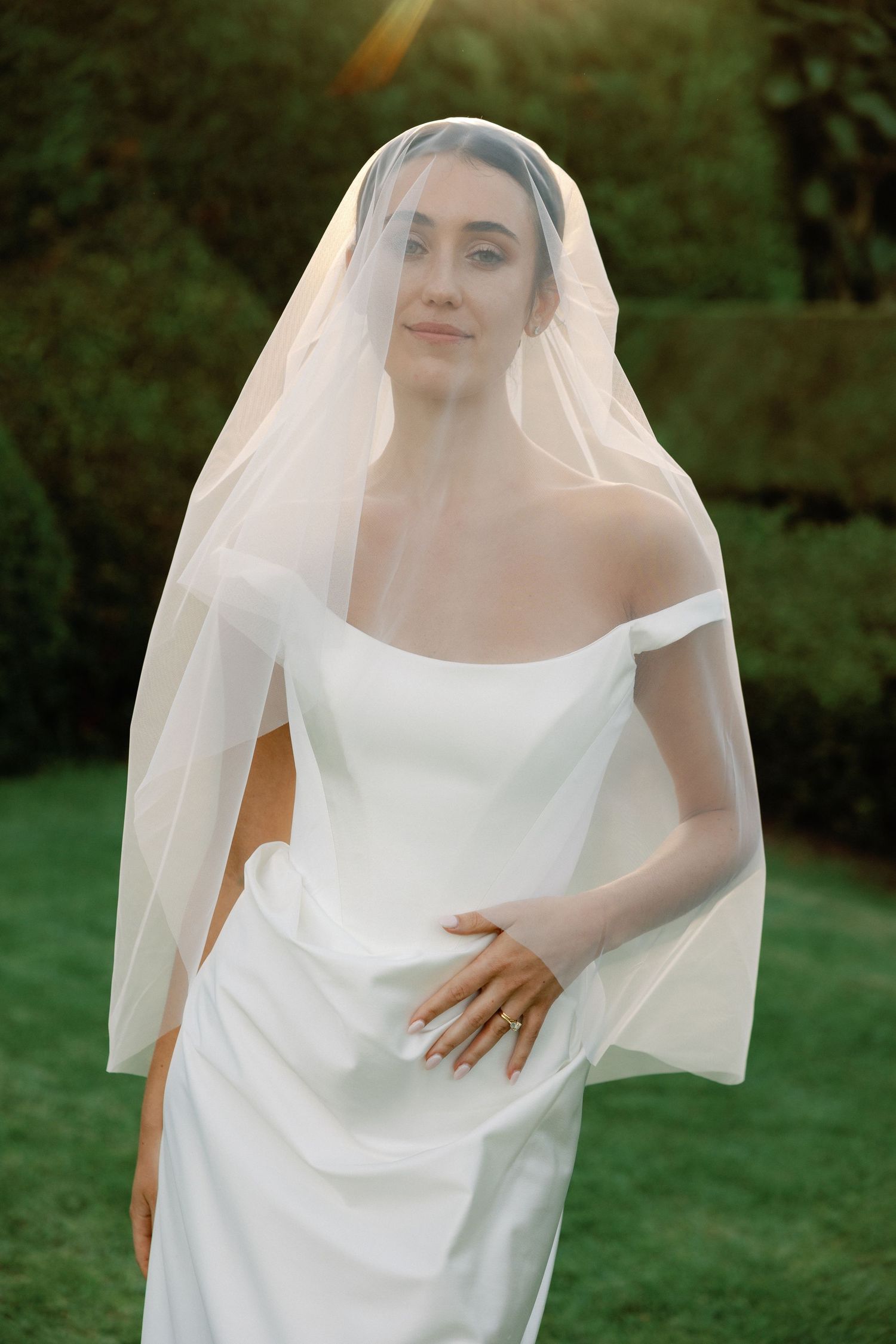 Bride in off-shoulder white gown and flowing veil poses outdoors amid lush green hedges in golden sunlight.