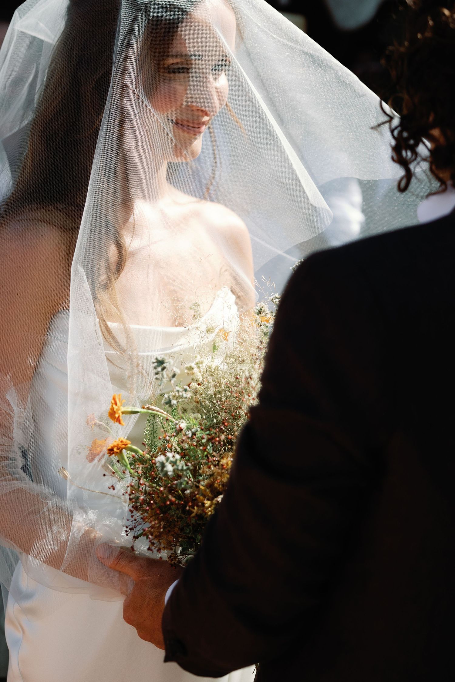 Bride in white gown and veil holds wildflower bouquet with orange blooms, facing groom in dark suit outdoors.