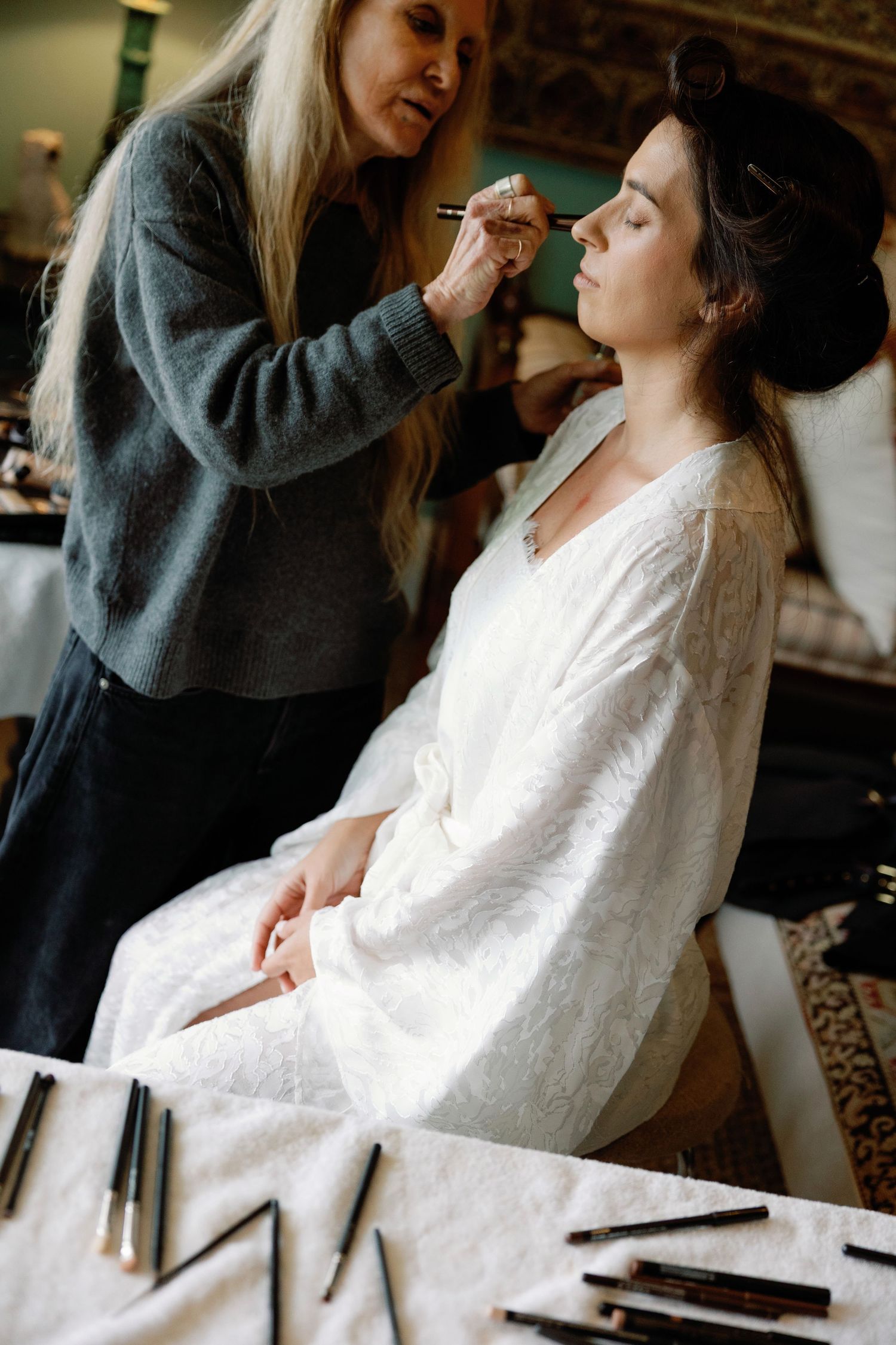 A makeup artist applies eye makeup to a bride in a white lace robe, with brushes laid out on a table nearby.