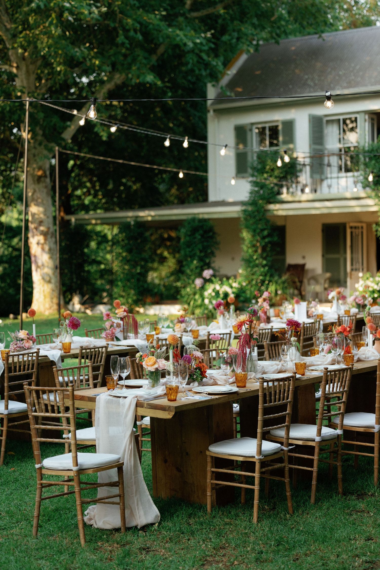 Elegant outdoor wedding reception with string lights, gold chiavari chairs, and colorful floral centerpieces on rustic wood tables.