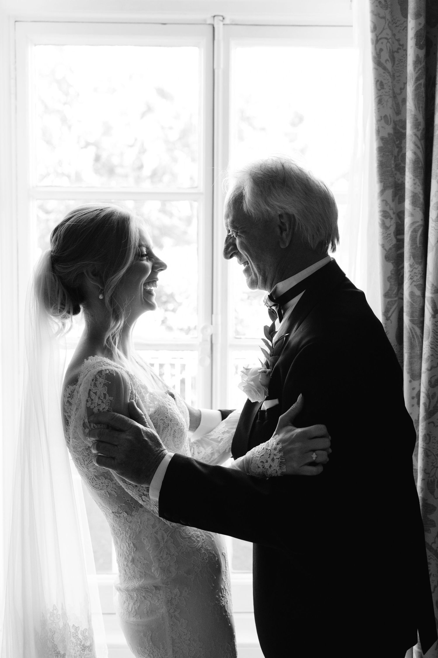 Black and white photo of a bride and elderly man sharing a tender dance moment by a sunlit window on wedding day.
