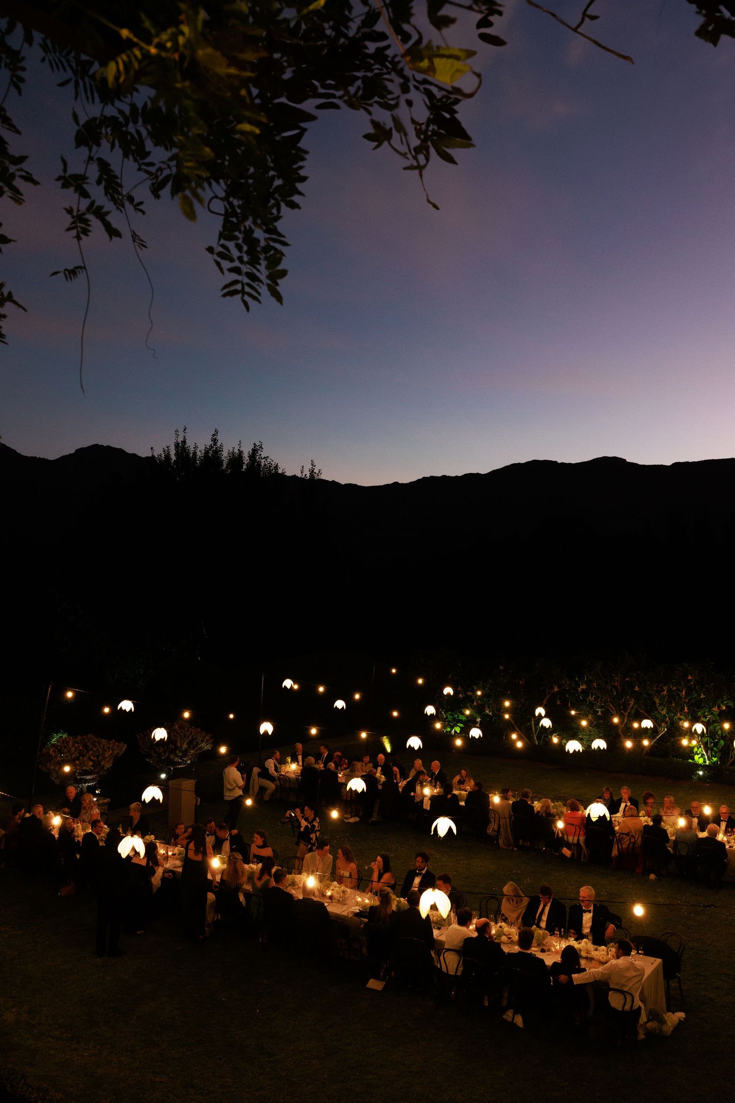 Outdoor evening wedding reception with glowing lanterns on tables, set against mountain silhouettes at dusk.