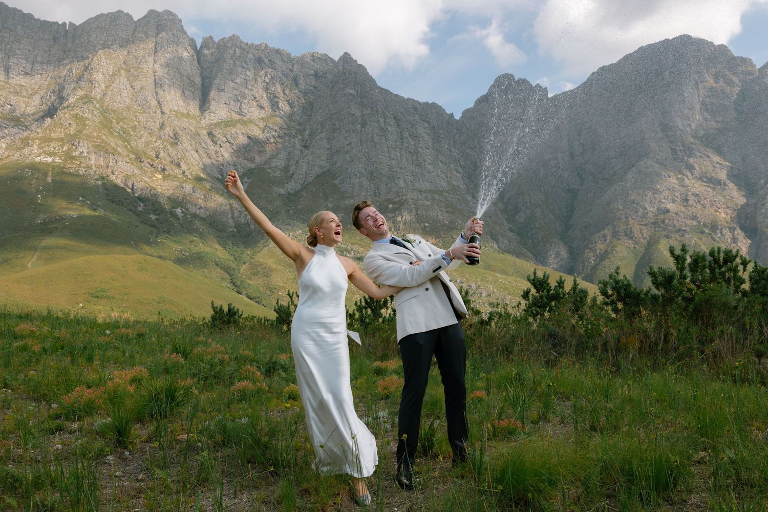 People celebrate with outstretched arms against a backdrop of majestic mountain peaks and green meadows.