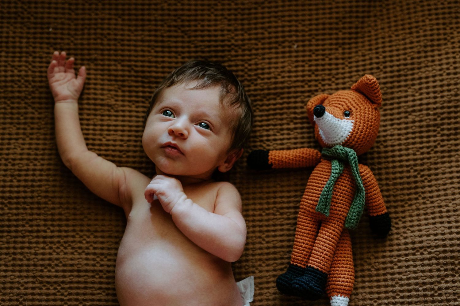A baby lying beside an orange knitted fox toy on a brown textured surface.