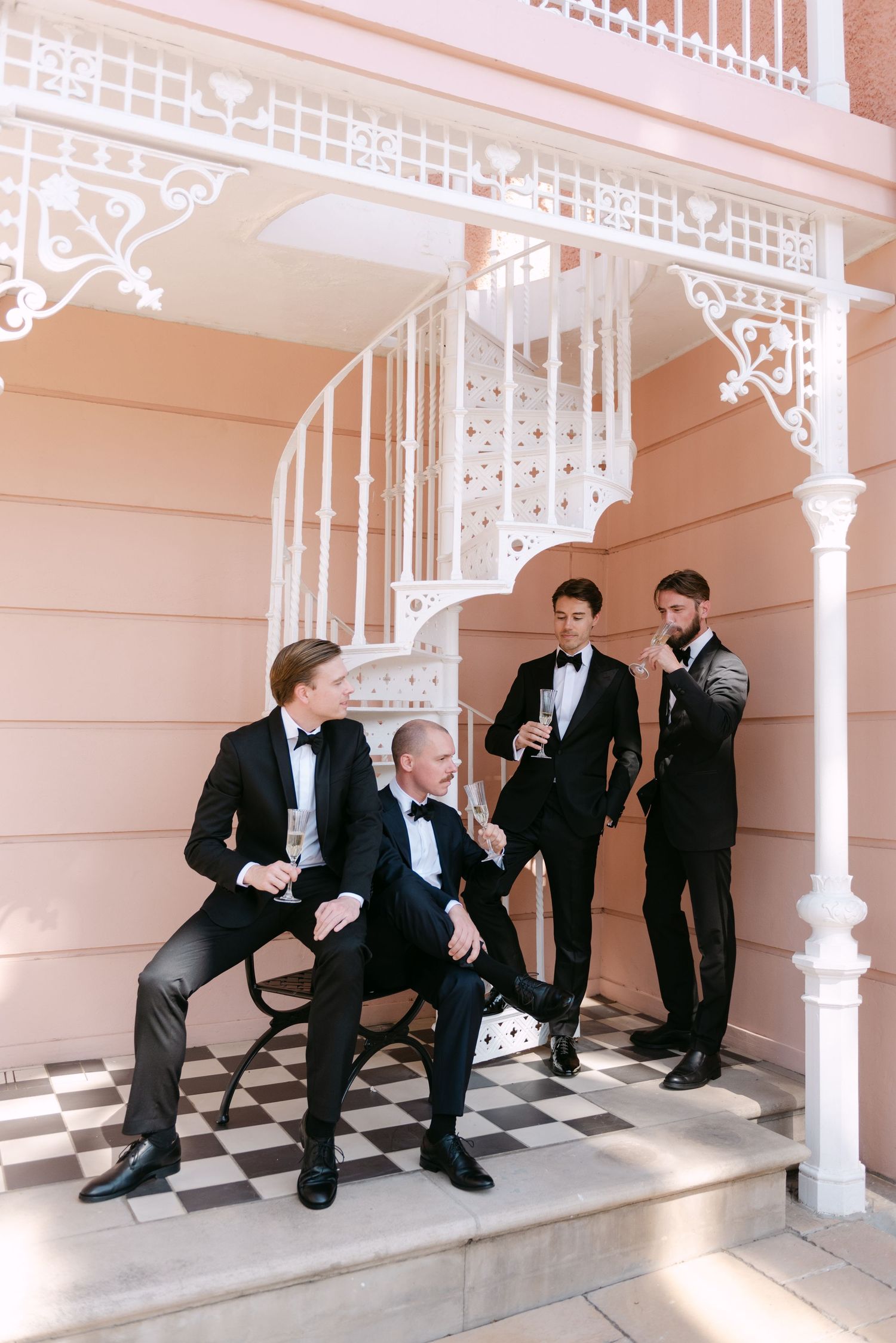 Four groomsmen in black tuxedos pose together on vintage white porch with ornate Victorian details.