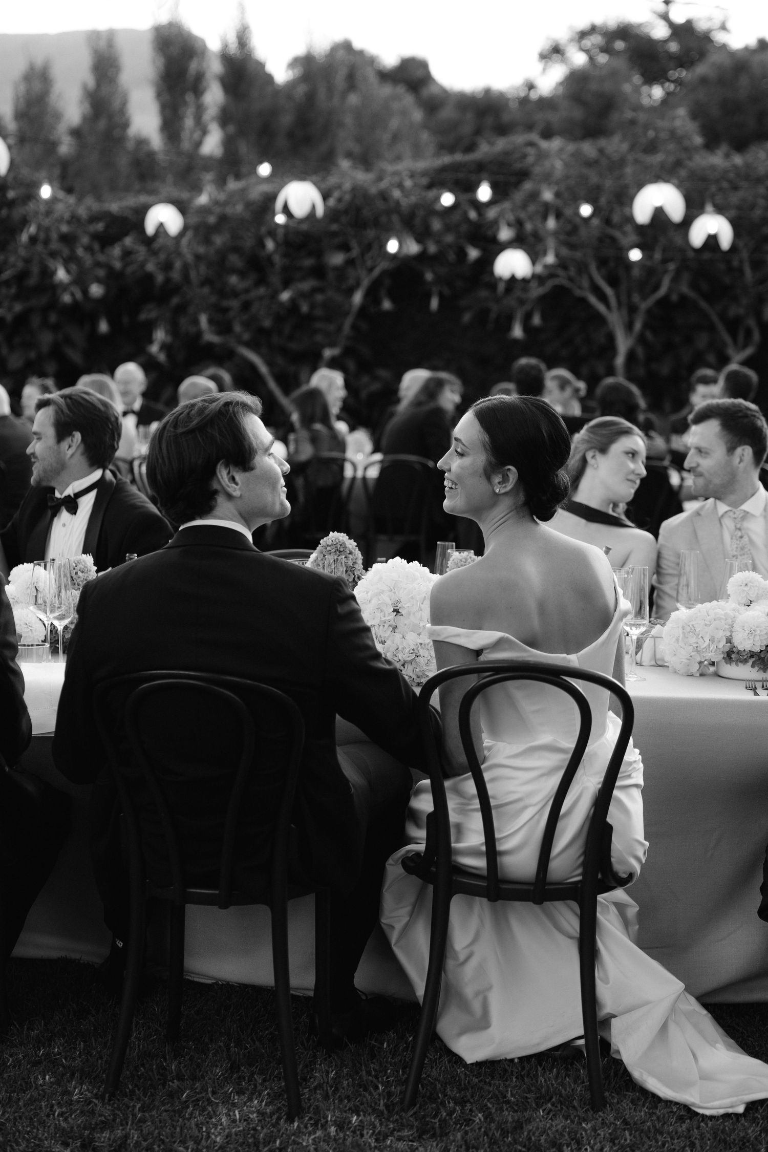 Black and white photo of bride and groom seated at outdoor wedding reception dinner table with guests and string lights.