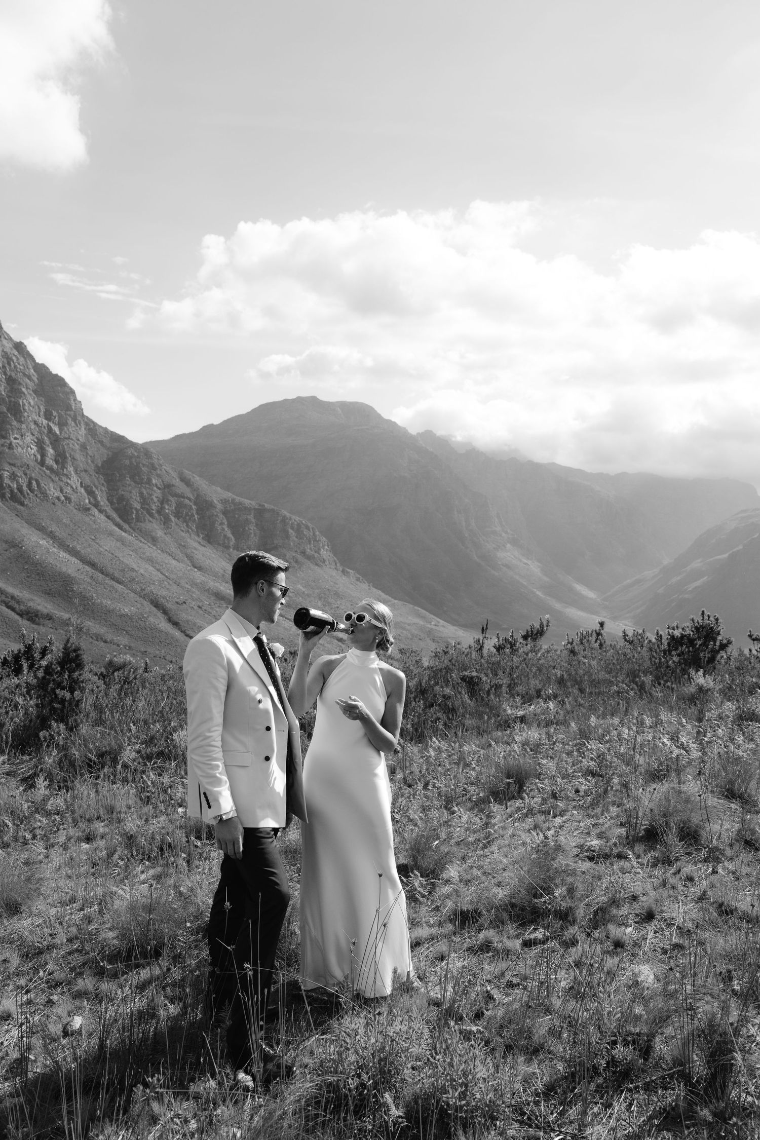 Two people walk through a scenic mountain landscape with dramatic peaks and clouds in black and white.