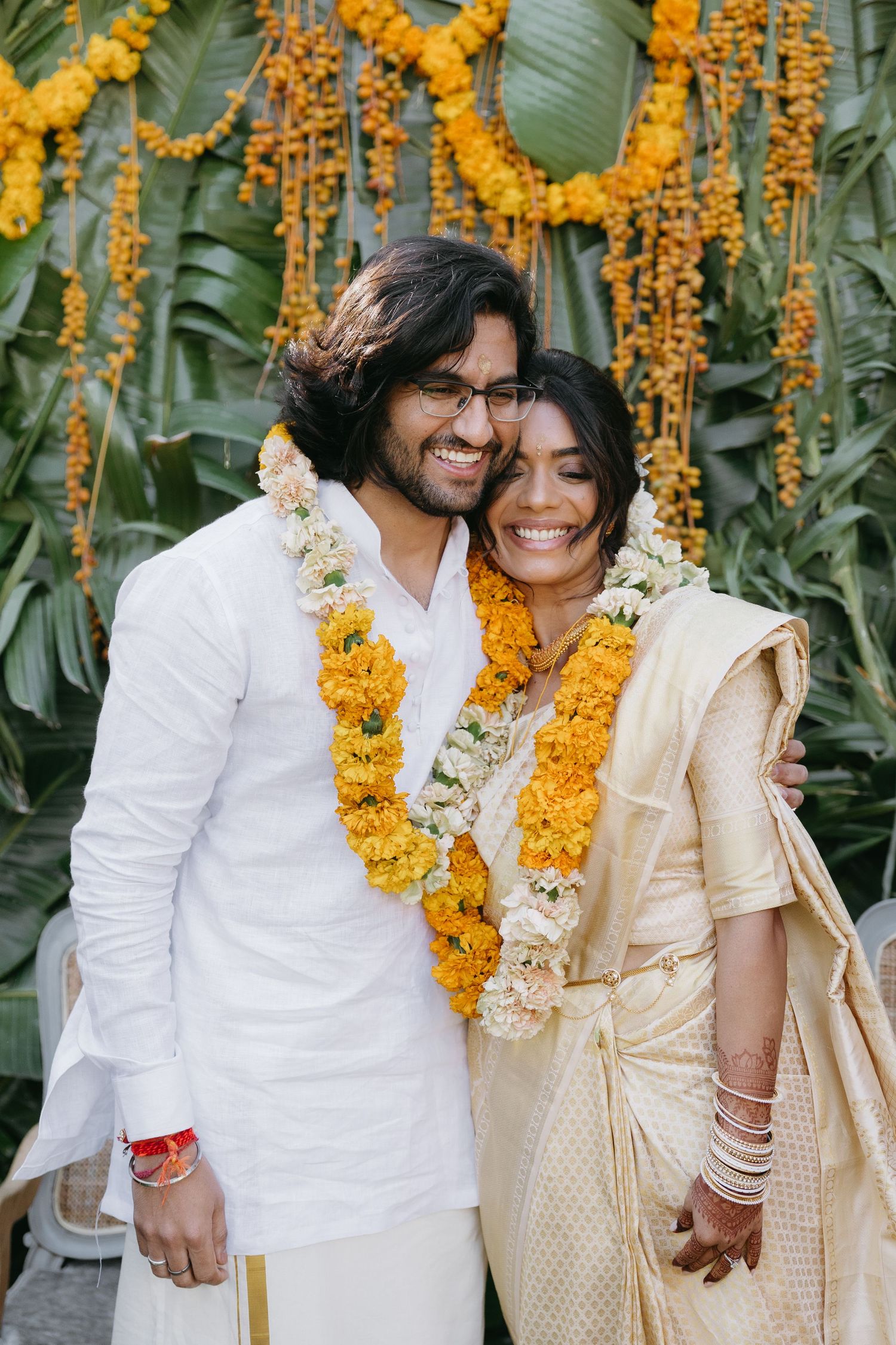 A traditional Indian wedding couple wearing marigold garlands against a tropical backdrop with yellow flowers.