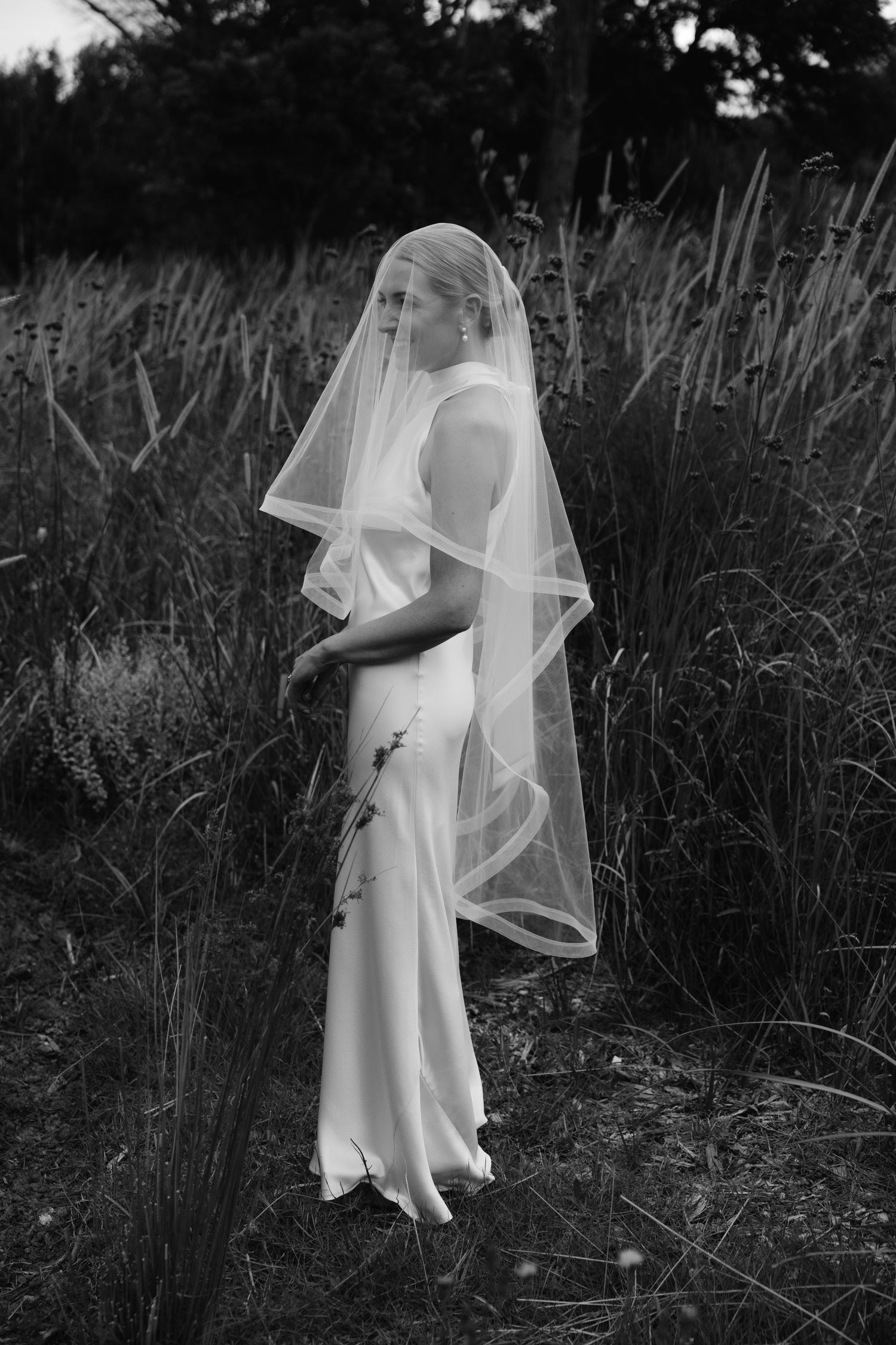 A person in a flowing white dress and veil stands in tall grass at dusk creating an ethereal silhouette.