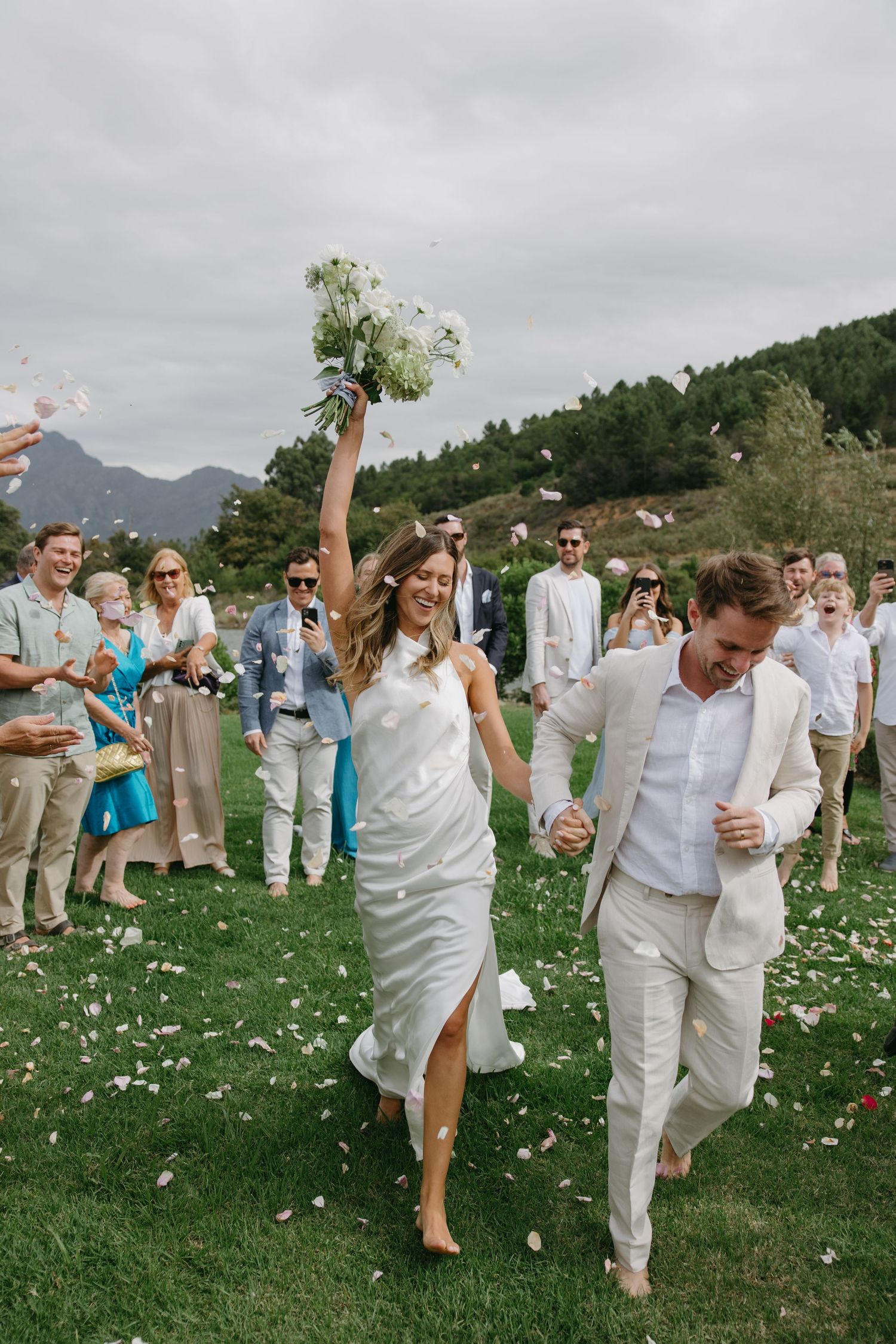 Wedding guests in white attire celebrate by dancing and throwing flower petals on a grassy hillside during an outdoor celebration.