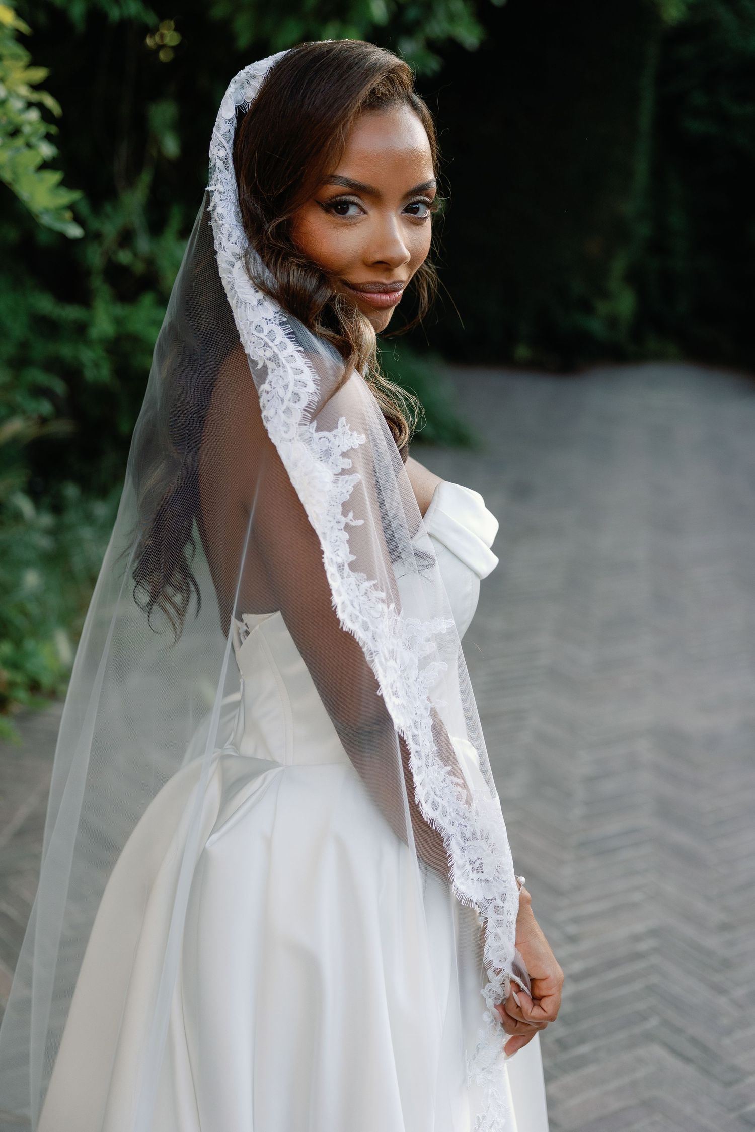 Elegant Black bride in white lace-trimmed veil and strapless gown poses outdoors against lush green hedges.