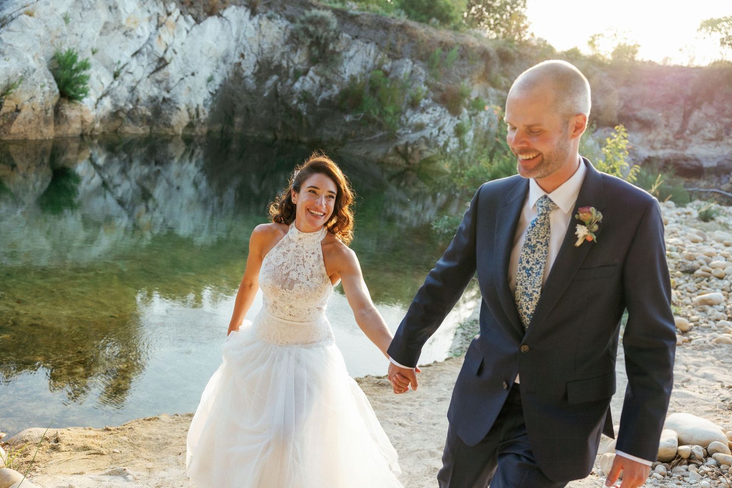 A couple in wedding attire holding hands and walking by a scenic pond with cliffs in the background during sunset.