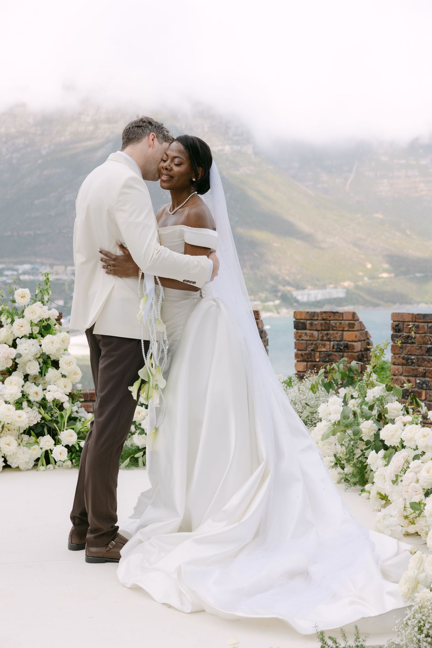 A romantic wedding couple embraces on a mountain terrace decorated with white floral arrangements and scenic views.