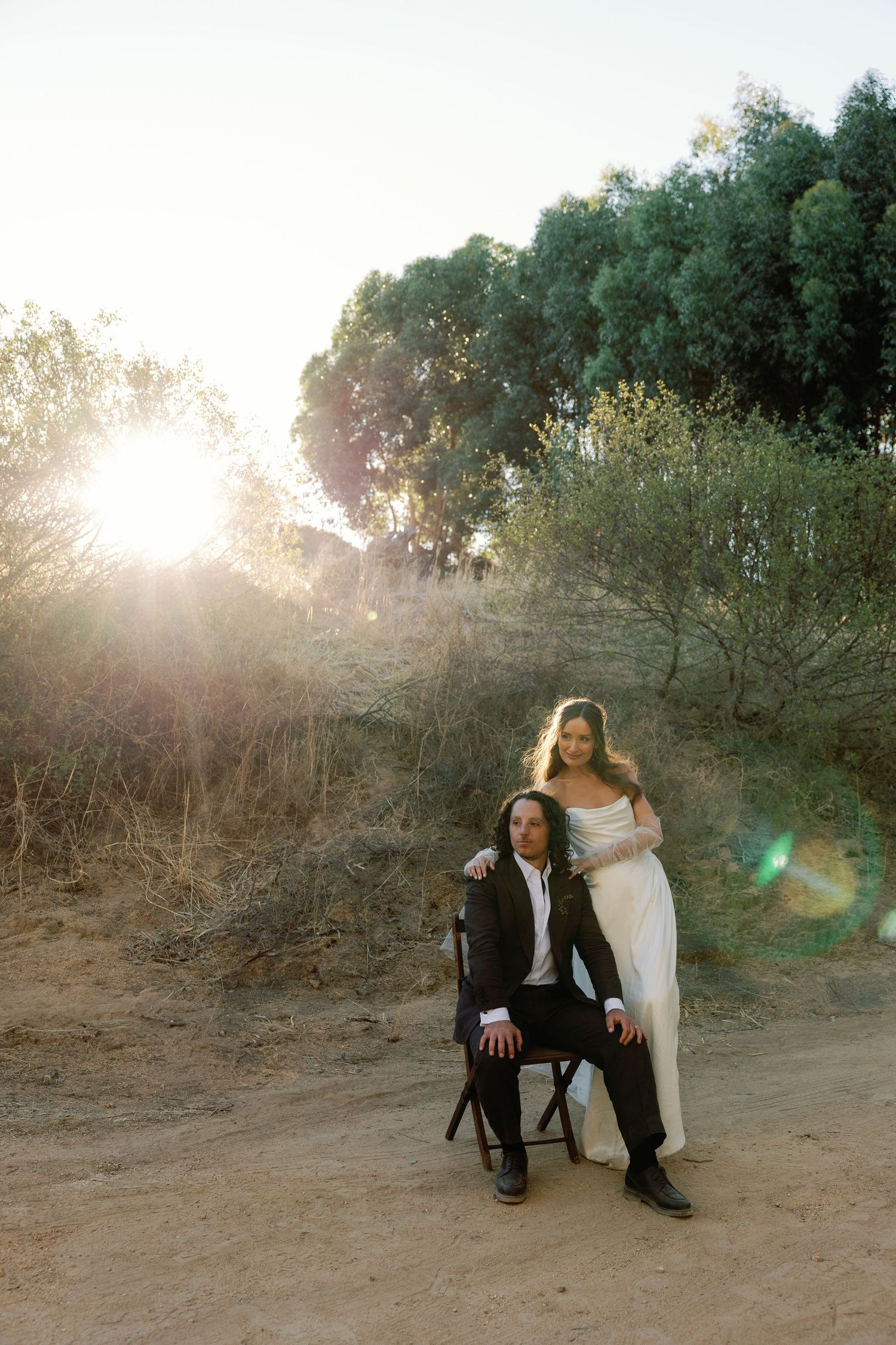 A wedding couple poses in a natural setting with golden sunlight streaming through trees in the background.