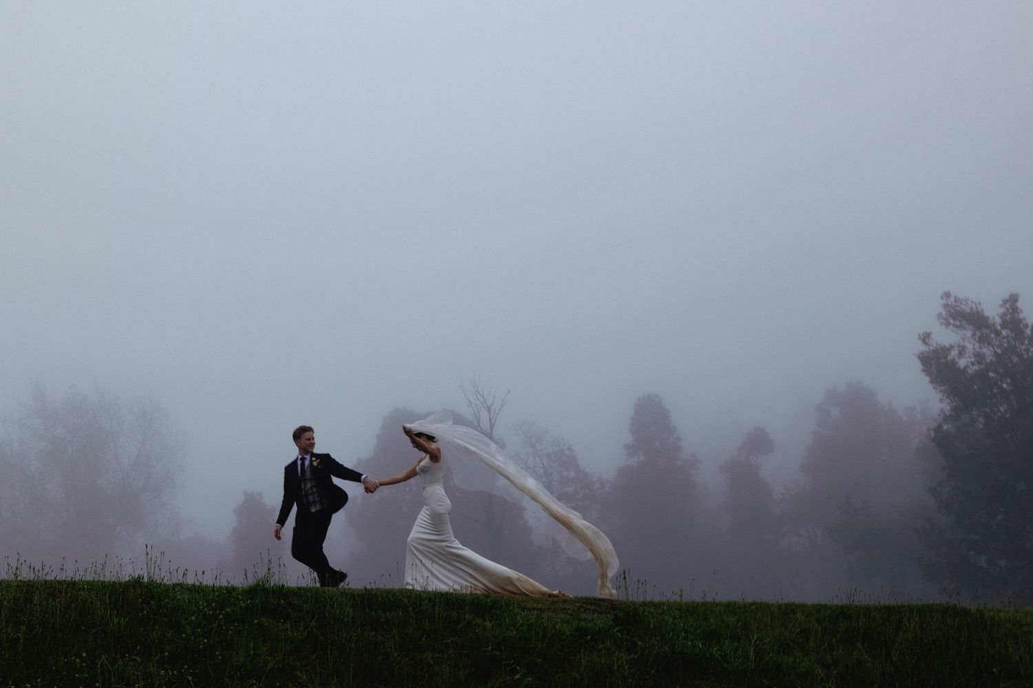 A couple poses on a misty hilltop as wind dramatically blows the bride's veil during a foggy outdoor photoshoot.