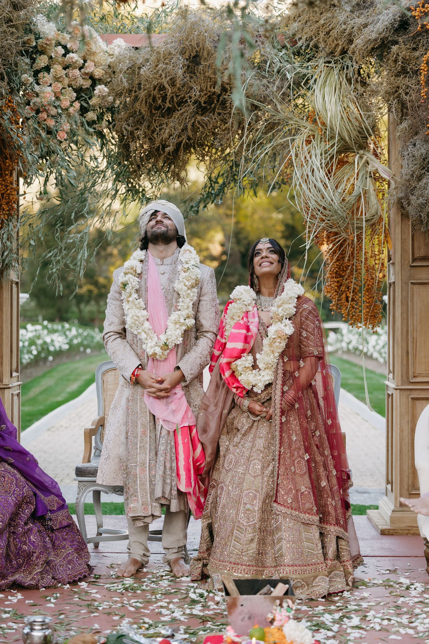 Outdoor Indian wedding photoshoot featuring traditional outfits and decor under trees with scattered flower petals.