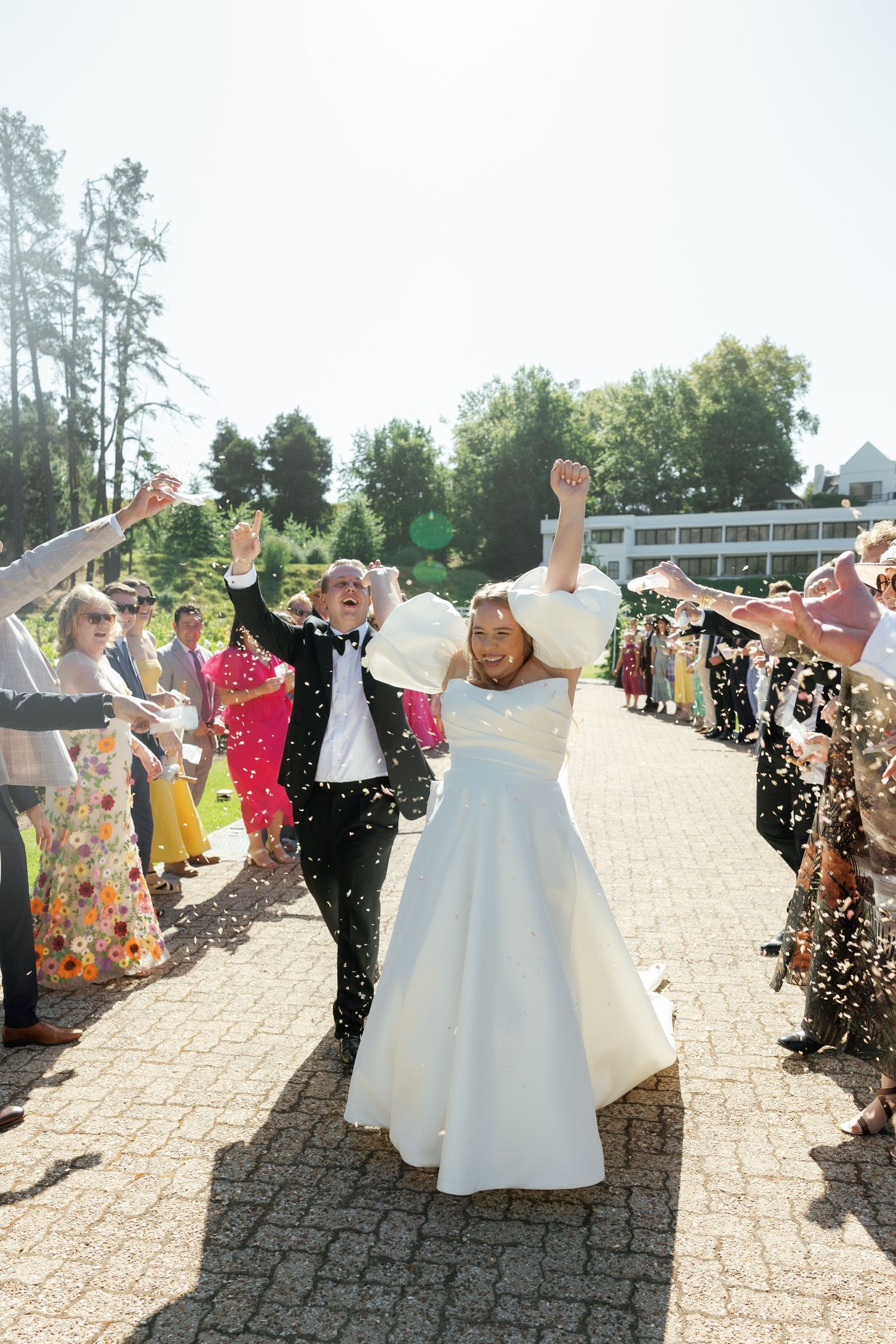Wedding guests shower newlyweds with confetti on stone patio as they walk through celebration line at outdoor venue.