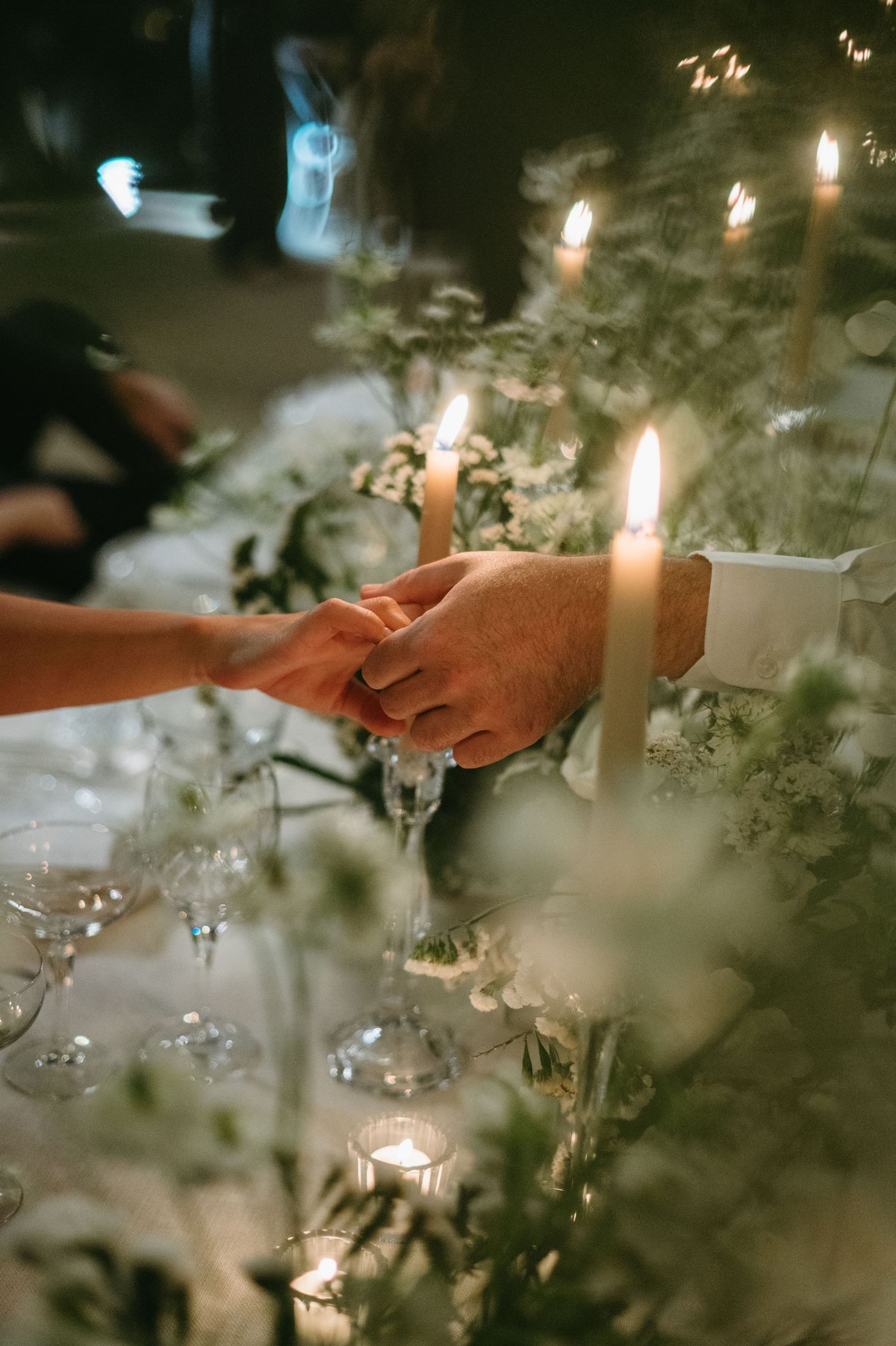 Hands reach across a candlelit table decorated with delicate floral arrangements and glassware at an elegant dinner setting.