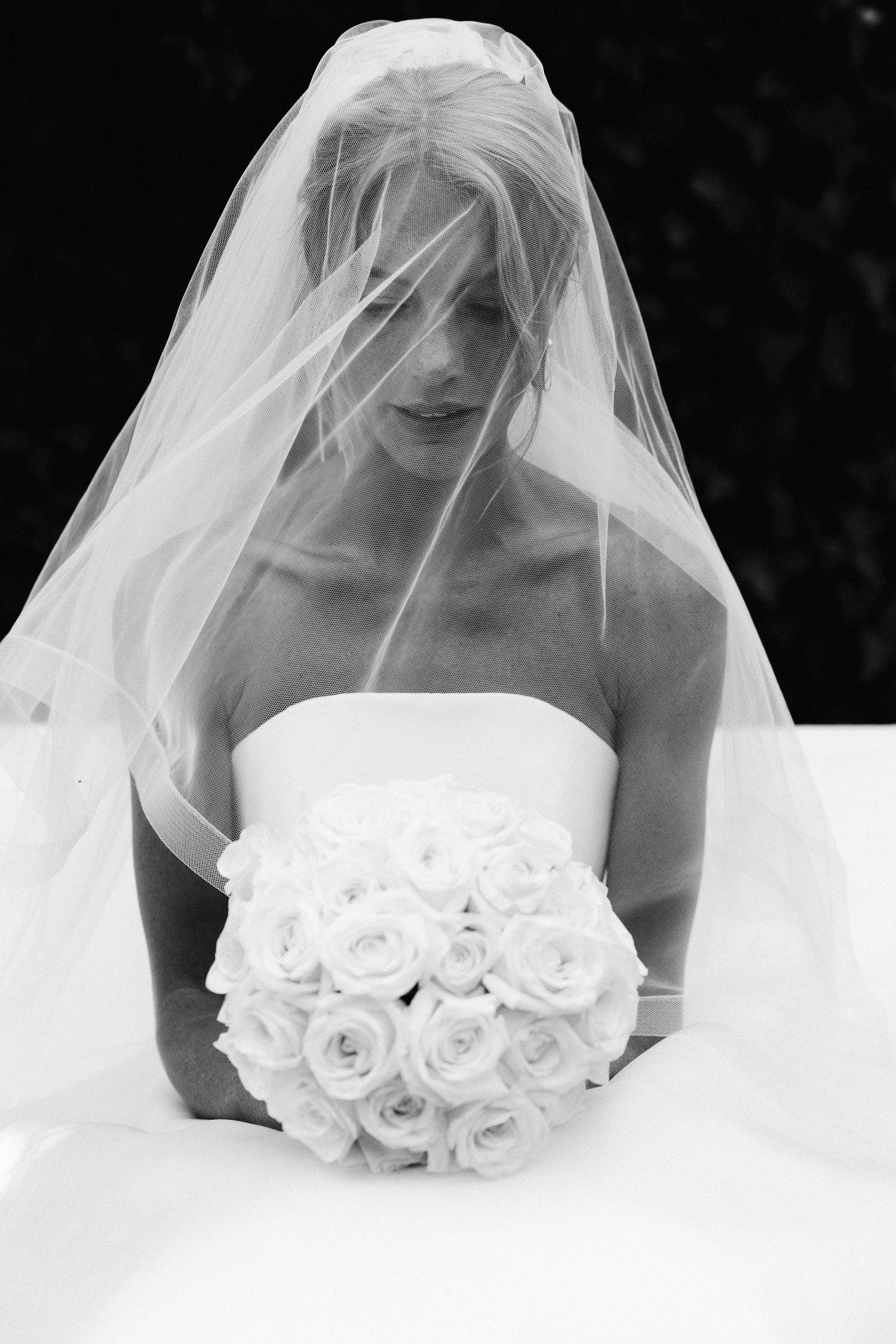Black and white photo of a bride in a strapless gown, veil draped over her face, holding a bouquet of white roses.