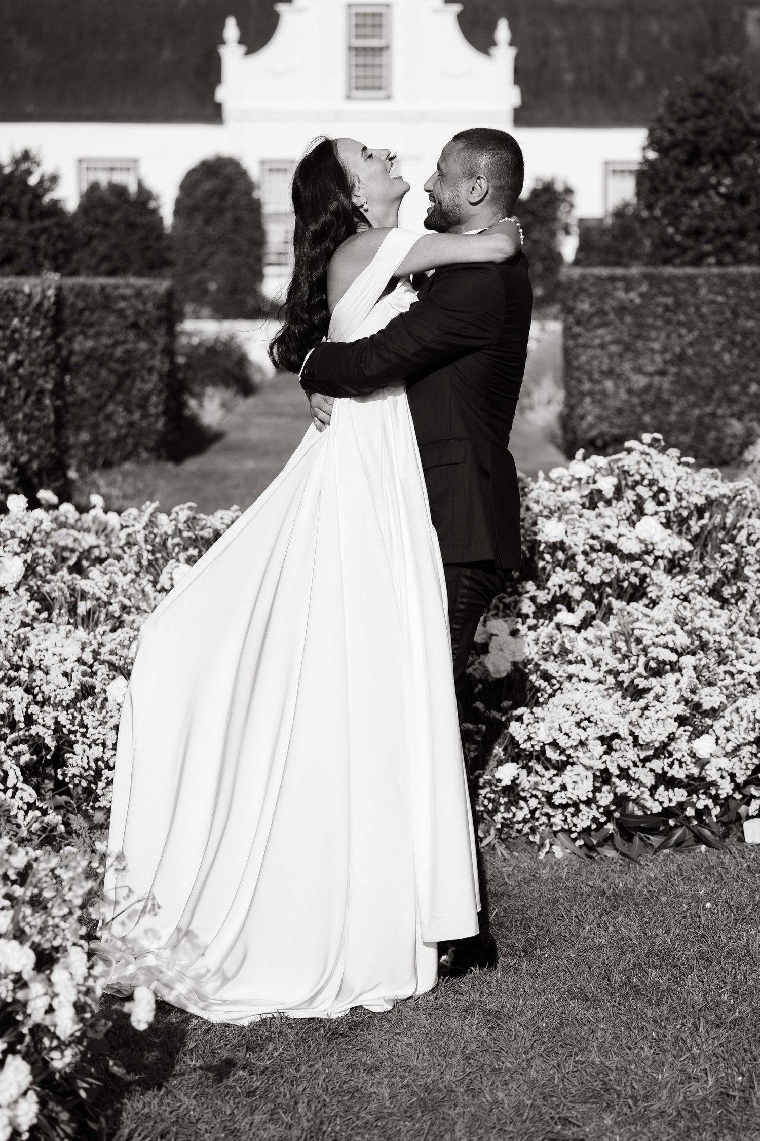 Bride and groom embrace in a romantic black and white wedding photo surrounded by blooming garden flowers.