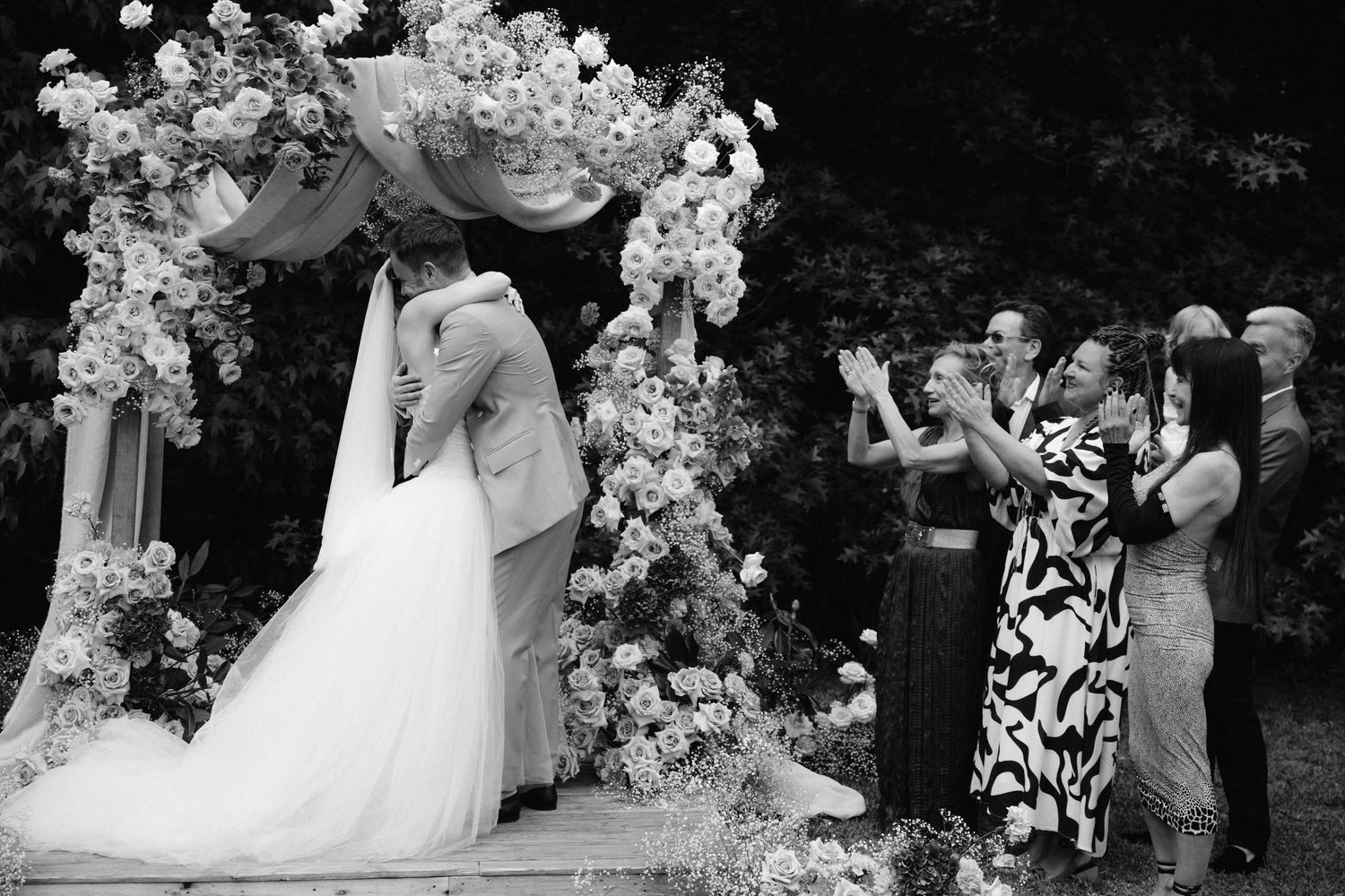 Black and white photo of a couple sharing their first kiss under a floral arch at an outdoor wedding ceremony.
