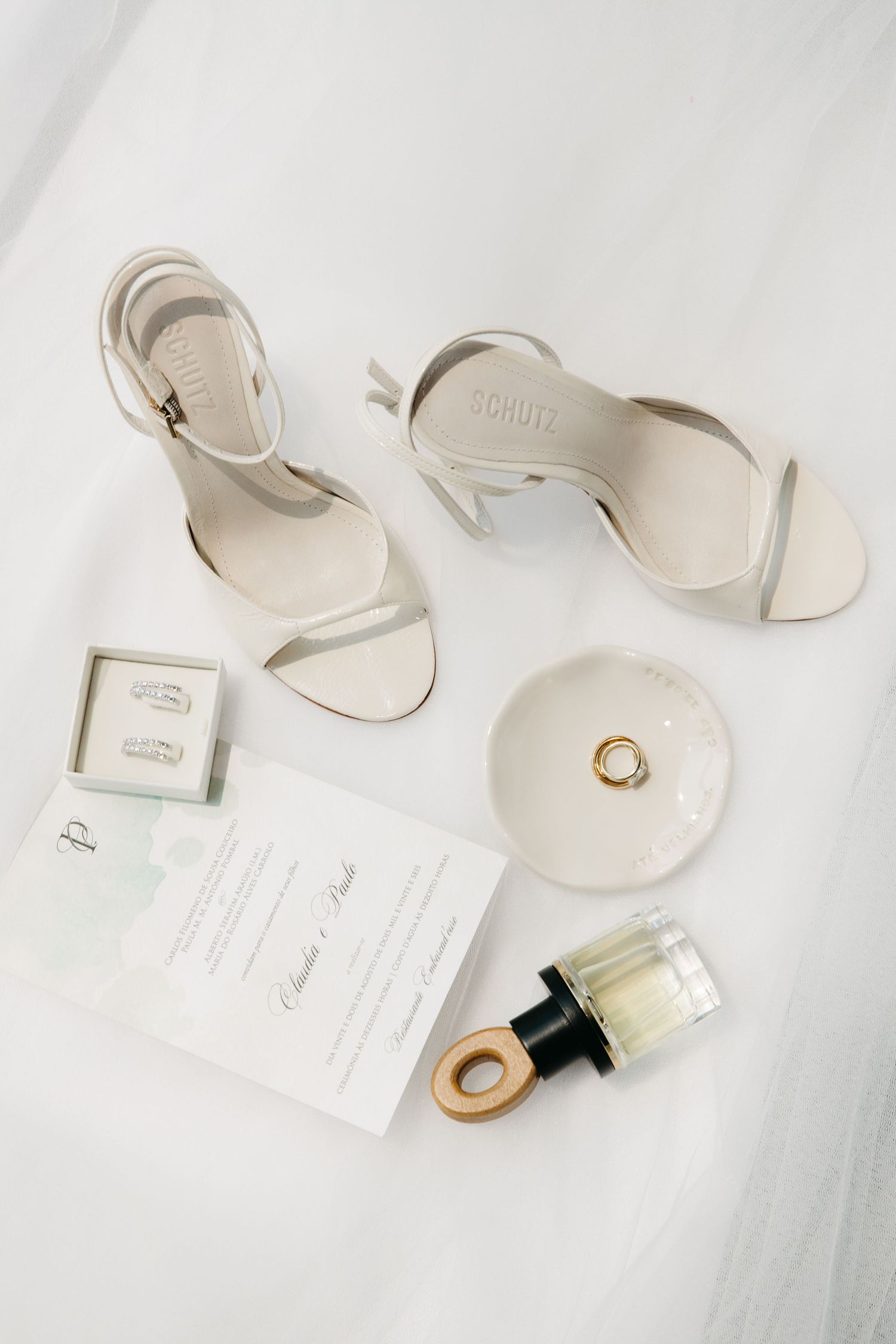 Flat lay of bridal accessories including white heels, gold rings, perfume bottle, and wedding invitation on white background.