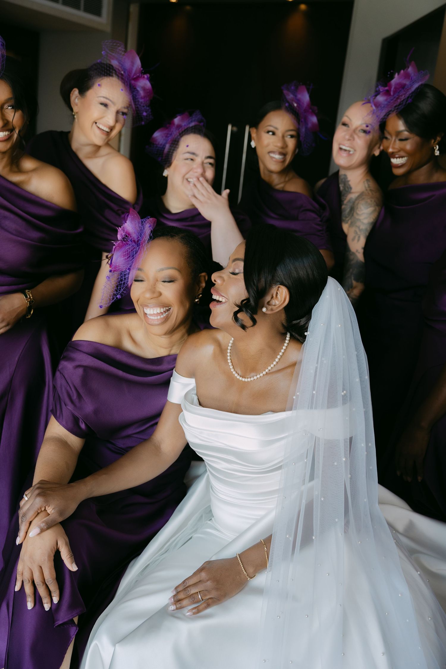 A joyful bride in a white wedding gown laughs with her bridesmaids wearing purple dresses during wedding celebrations.