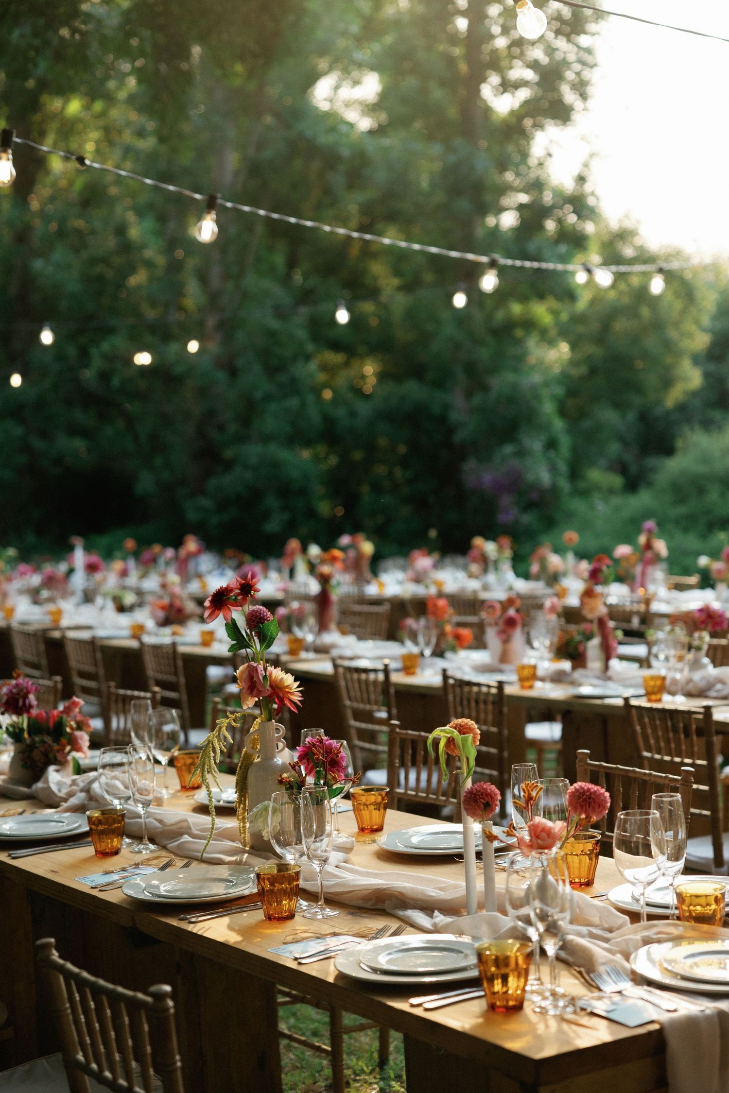 Elegant outdoor wedding reception tables set with amber glassware and floral centerpieces under string lights at dusk.