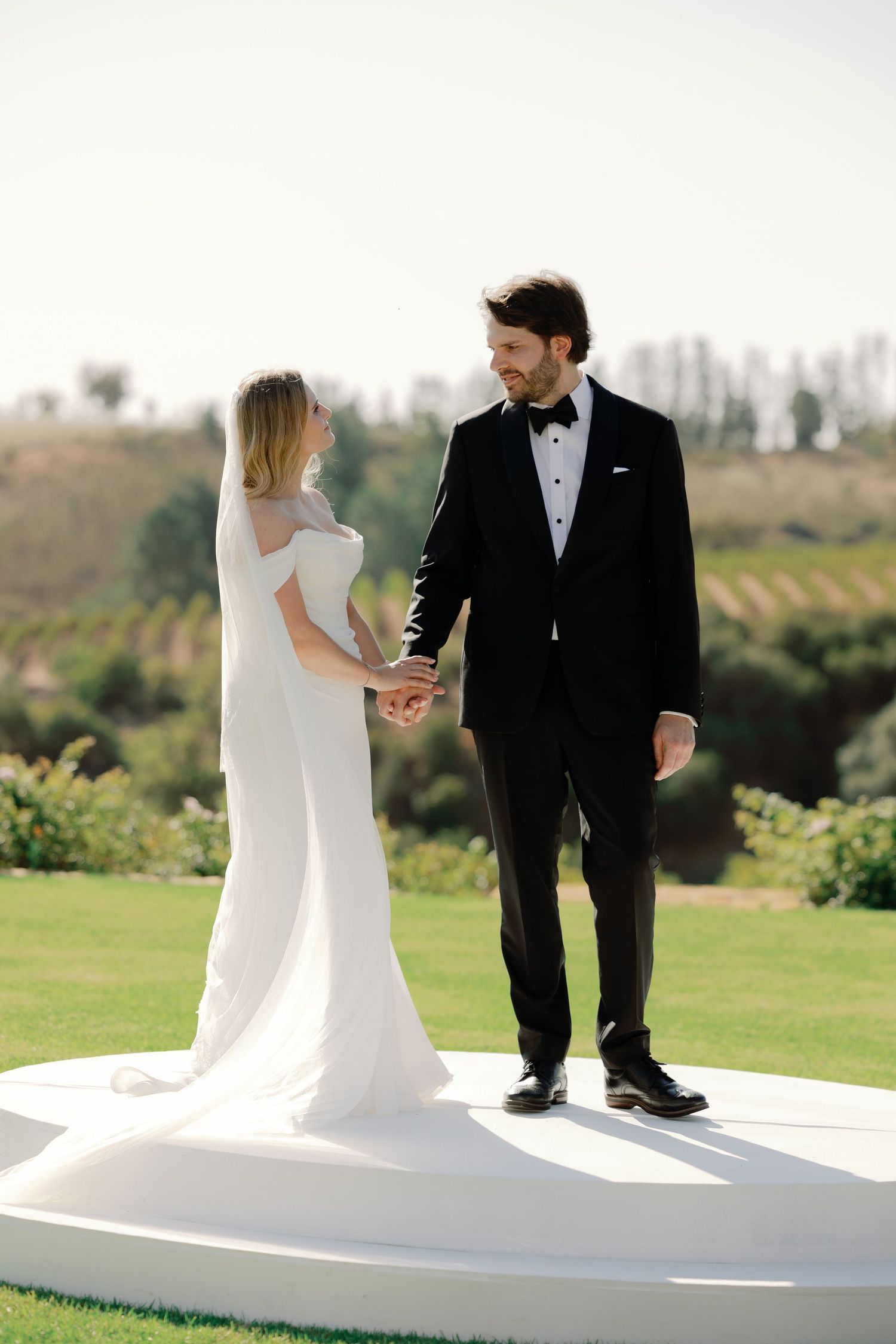 Bride and groom hold hands on white platform at outdoor wedding ceremony with green vineyard backdrop.