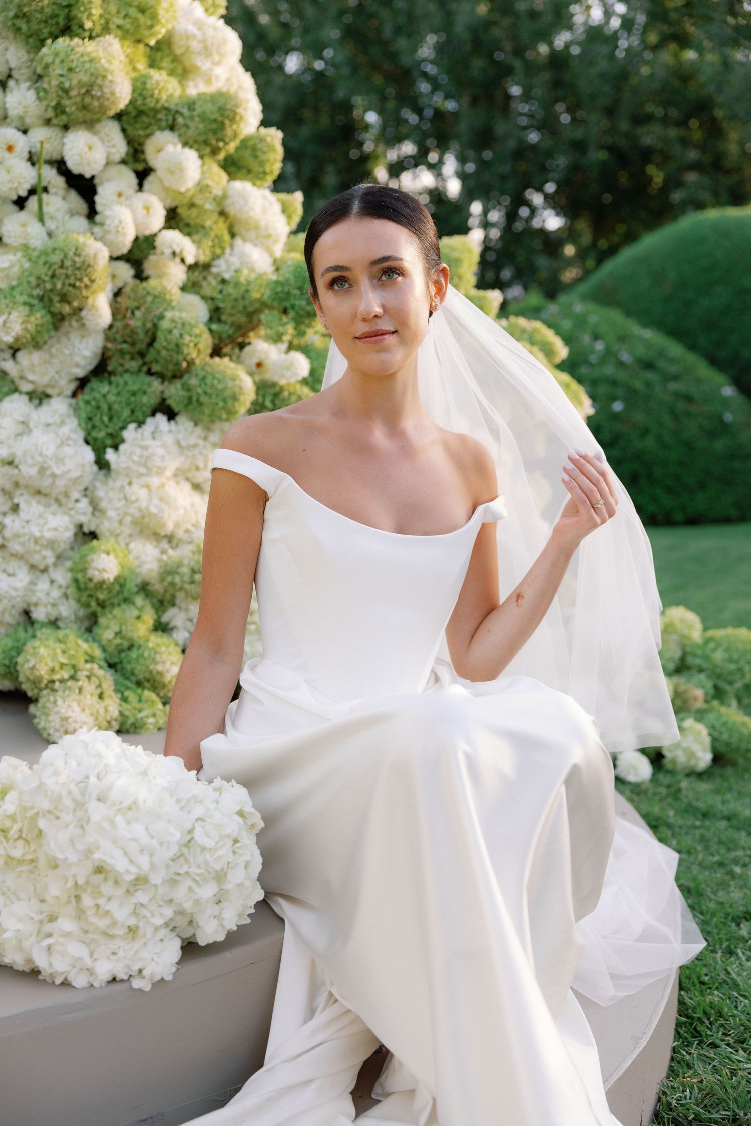 Elegant bride in off-shoulder white gown and veil sits among lush hydrangea arrangements in a garden setting.