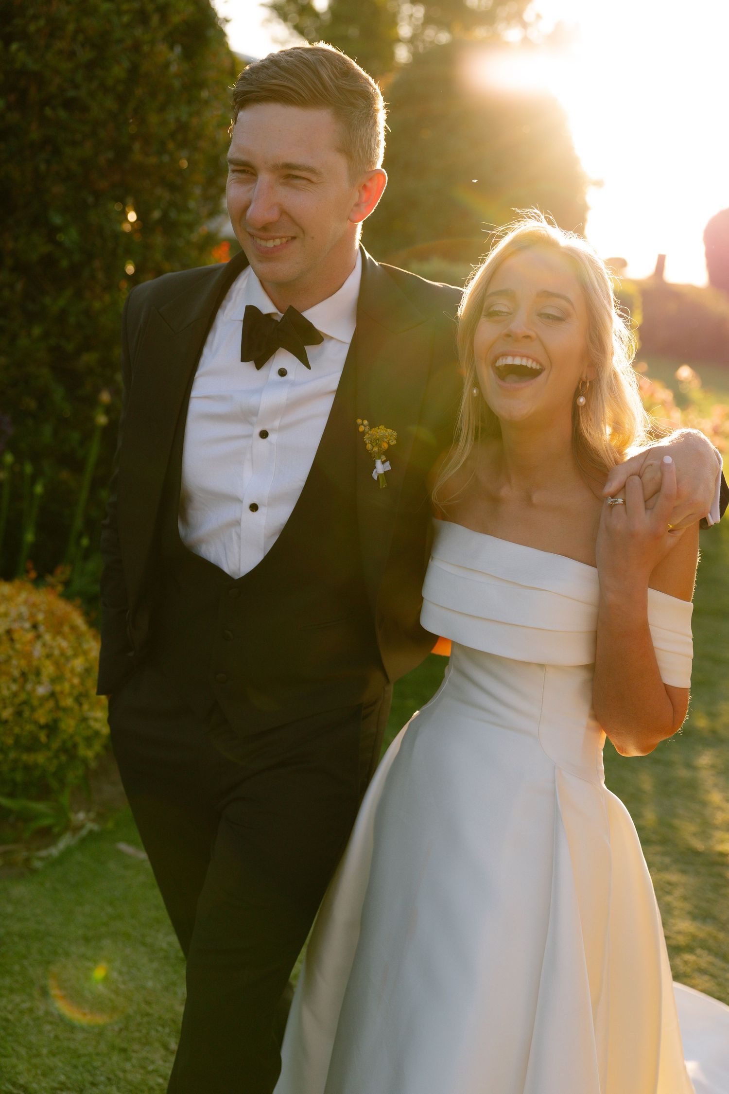 A happy couple laughs together during golden hour on their wedding day, wearing classic formal black and white attire.